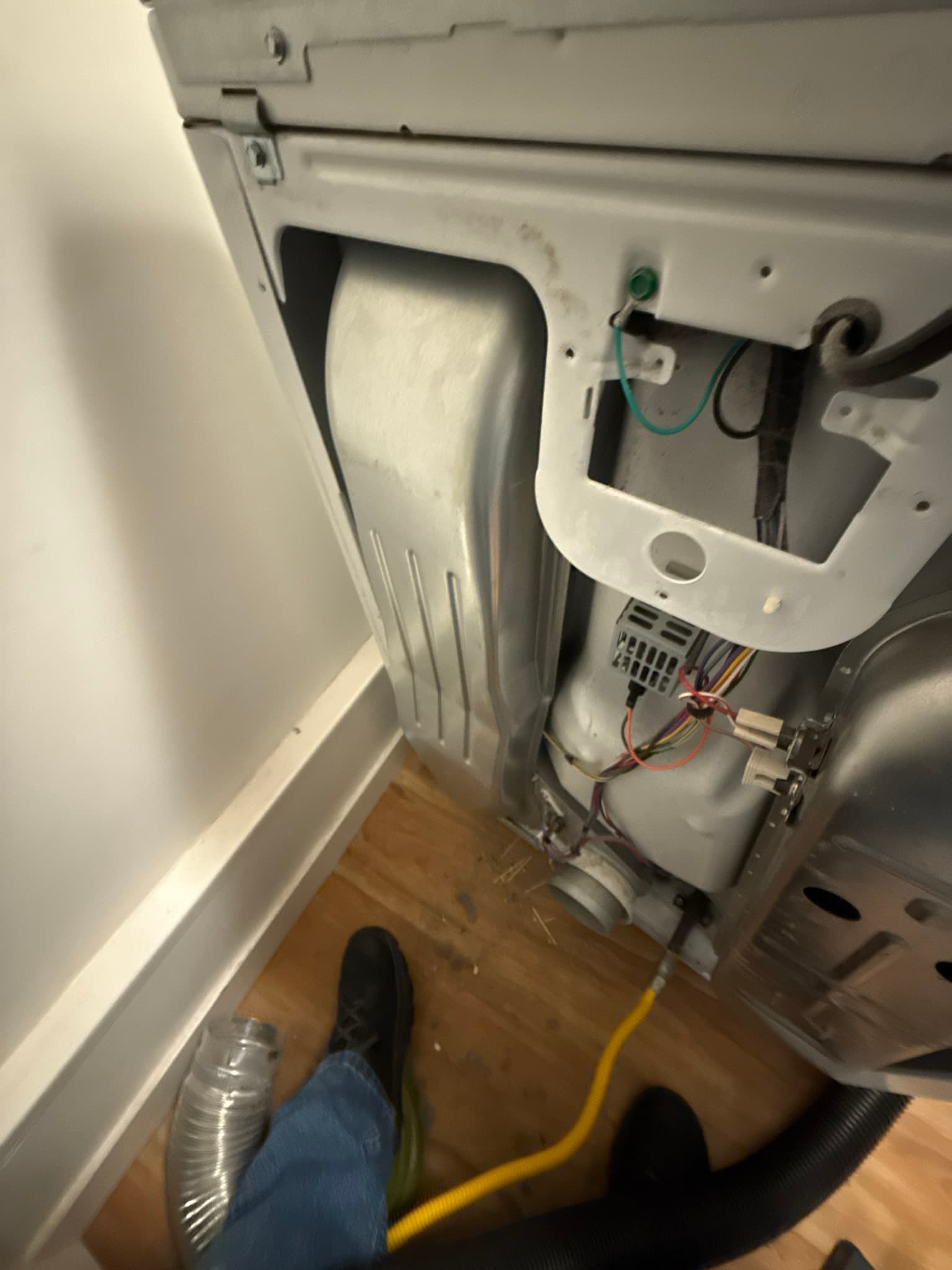 Interior of an electric dryer with the rear access panel removed, showing the dryer drum on the left, blower wheel on the right, wiring including a teal-colored wire and red and black power connections, and a yellow flexible dryer vent hose at the bottom