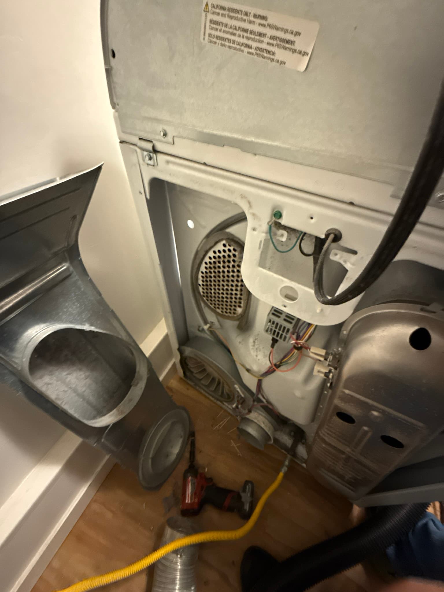 Rear panel of a residential electric dryer with the access cover removed, showing the exhaust duct connection point, internal heating components, and a yellow flexible duct at the bottom; dust and lint accumulation are visible on interior surfaces near the exhaust outlet