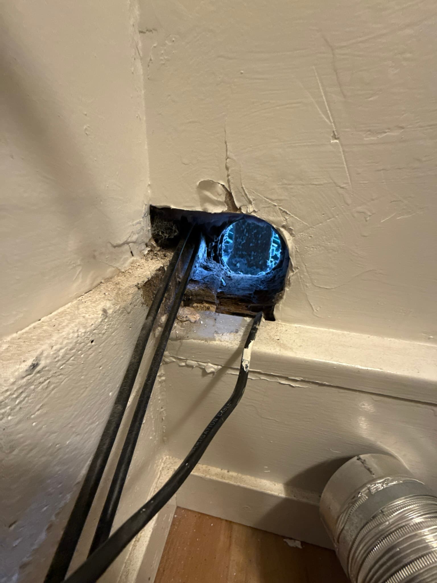 Dryer exhaust duct penetration through an interior wall showing a corroded blue metal elbow fitting heavily clogged with lint and debris, with gaps visible around the duct where it passes through the drywall