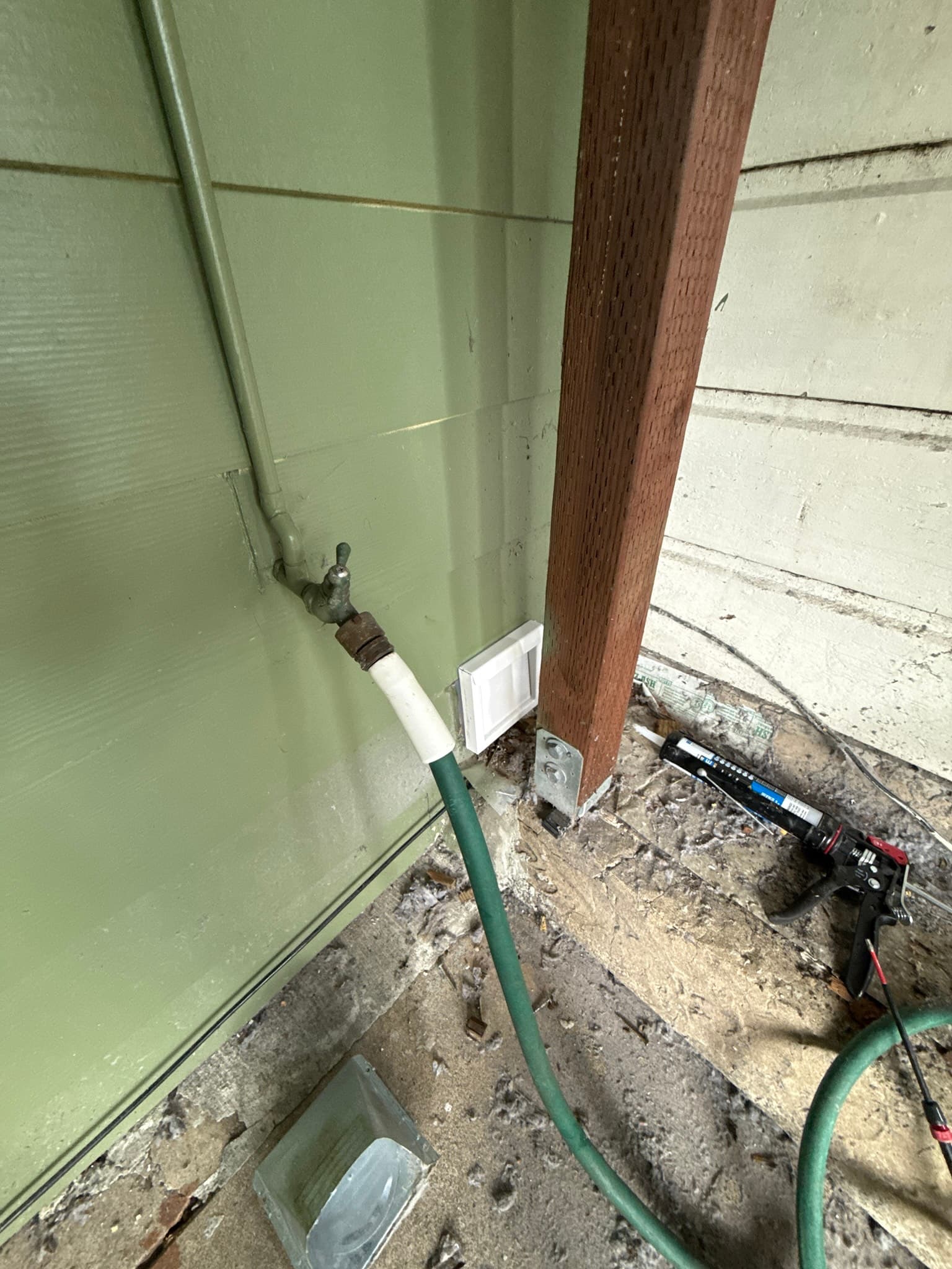 Dryer exhaust vent exiting through a foundation wall, with a green flexible duct connected to a corroded white rigid transition fitting at the wall penetration, surrounded by dirt and debris accumulation on the foundation surface