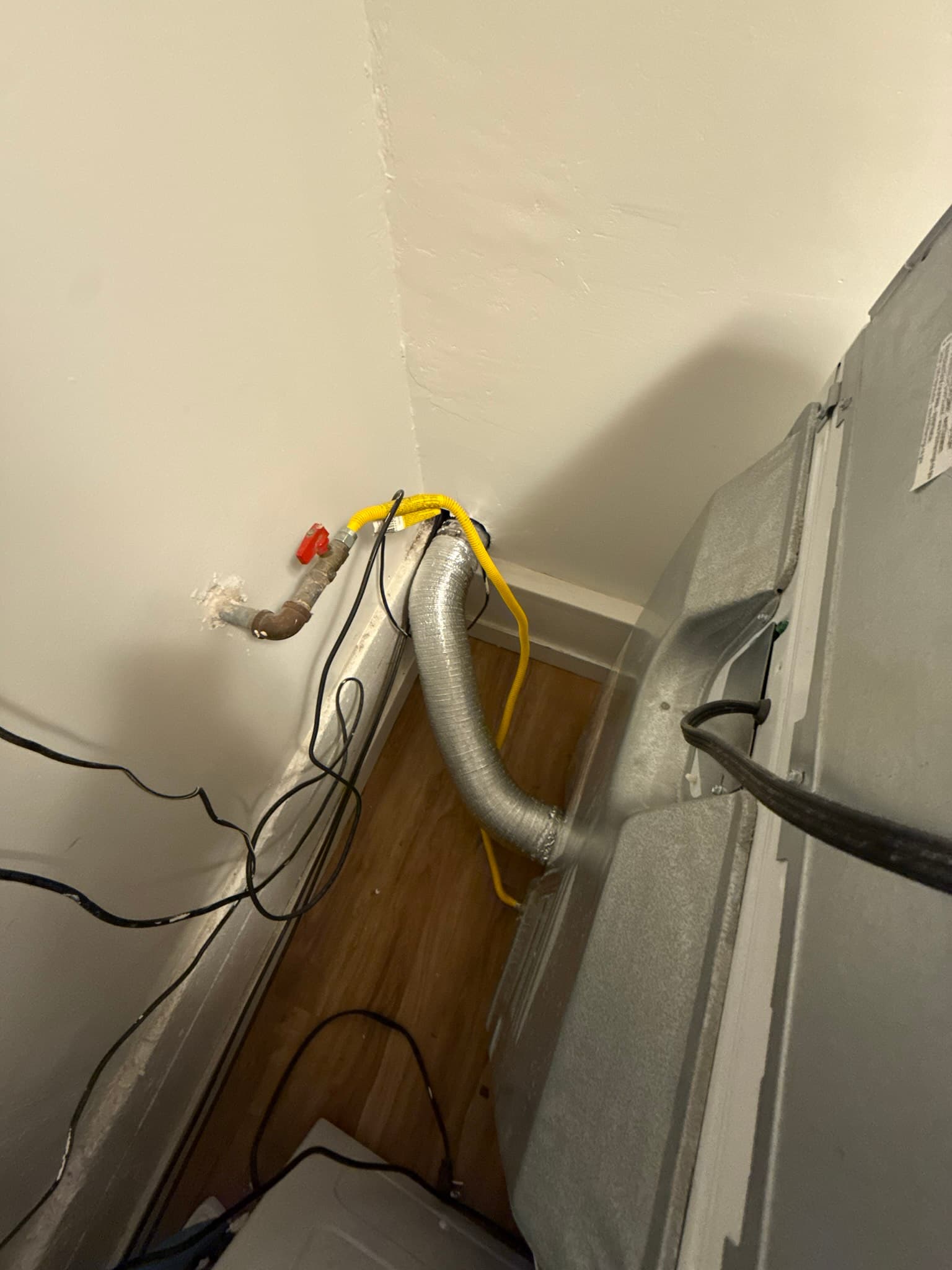 Dryer vent installation showing a silver flexible metal duct secured with yellow and black strapping, connected to a wall-mounted dryer unit, with a red manual damper visible near the wall penetration and multiple black power cords present