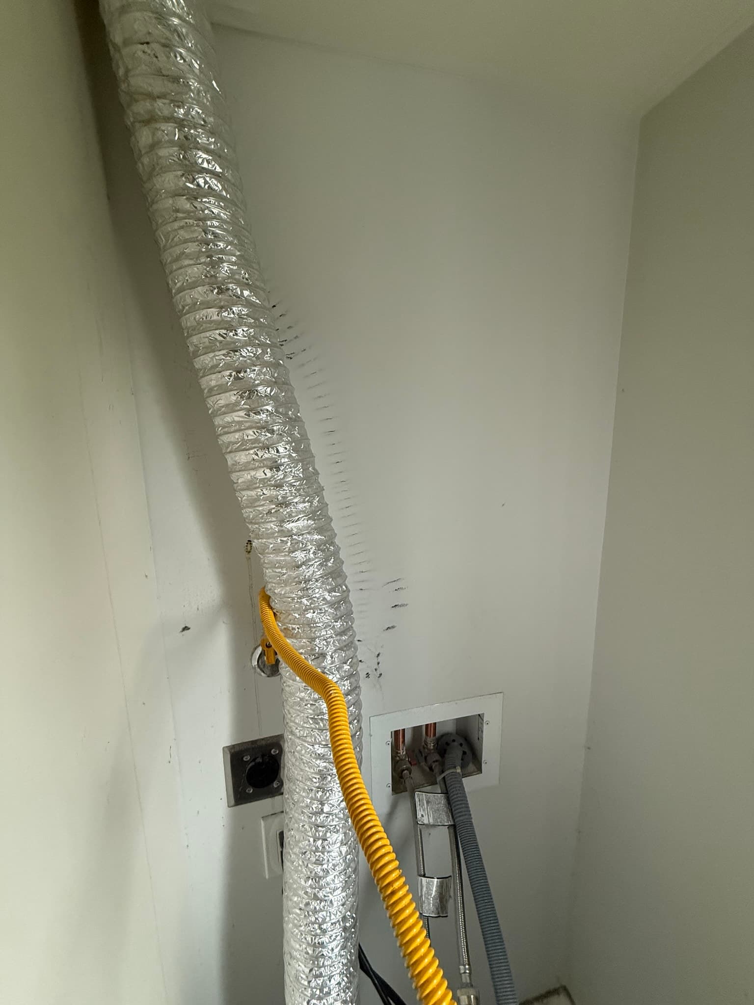 Existing flexible foil dryer vent duct running vertically along an interior wall, wrapped in reflective insulation, with a yellow conduit coiled at the base and no mechanical clamps or support straps securing the duct along its run