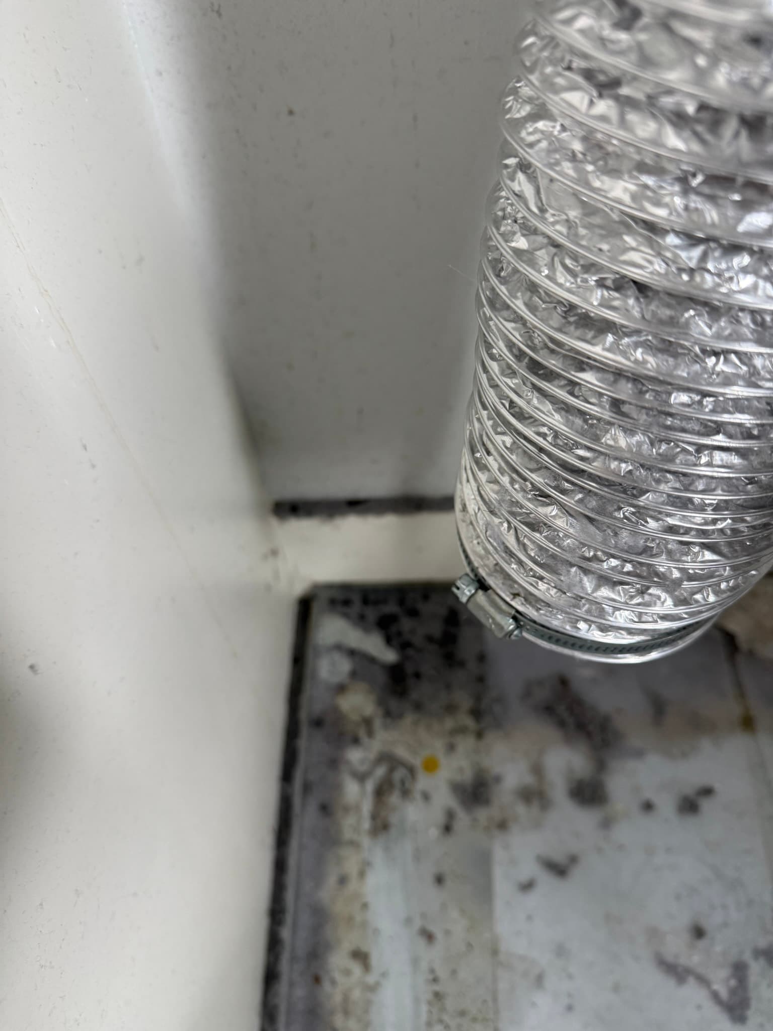 Flexible aluminum dryer vent duct connecting to a transition fitting, with dust and debris visible on the floor surface around the base, suggesting lint escape at the junction