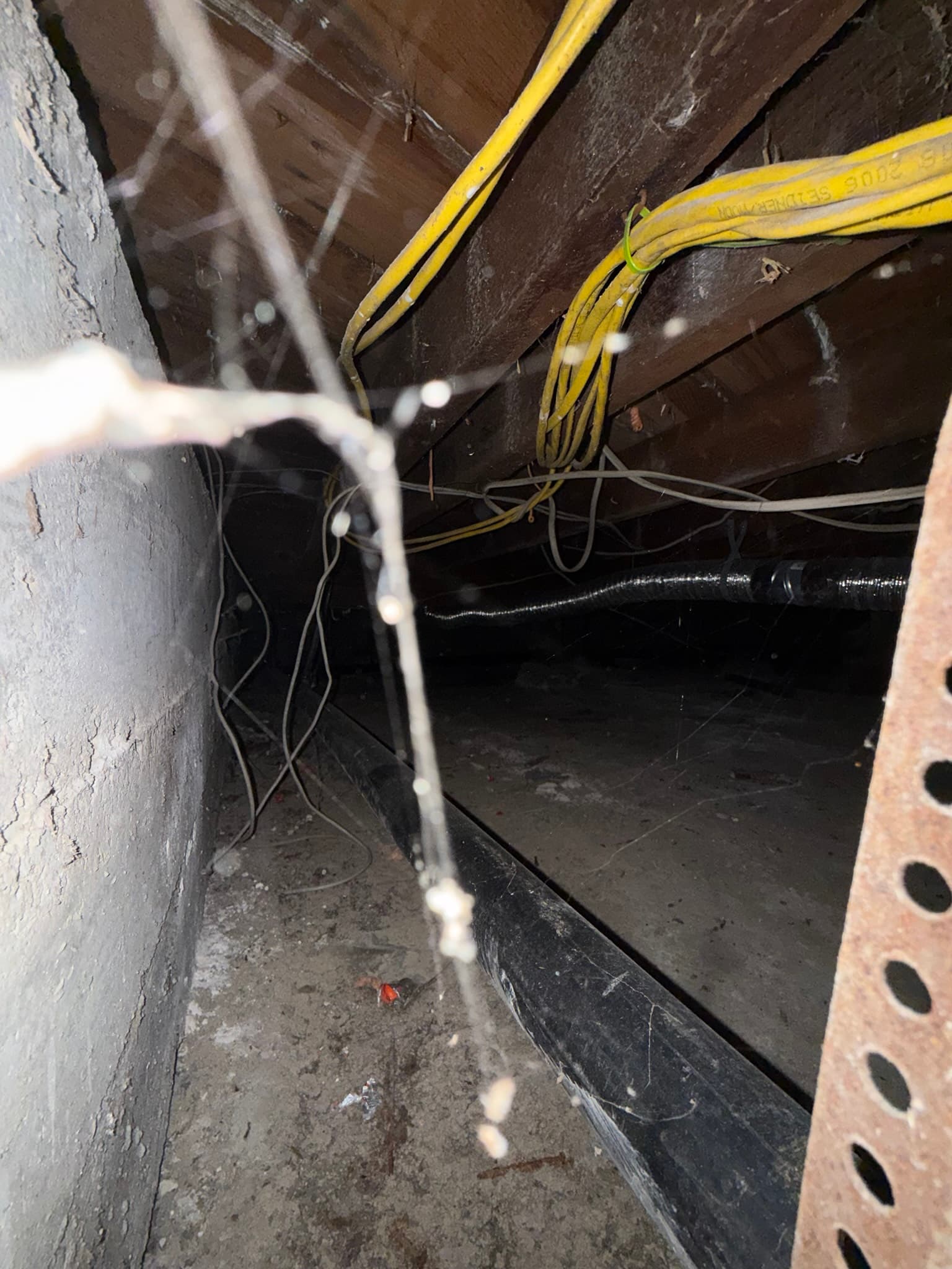 Attic or crawl space framing area with white lint deposits visible on dark wood joists and surfaces, a black flexible duct coated in accumulated lint running across the lower portion of the frame, and yellow electrical cables routed overhead