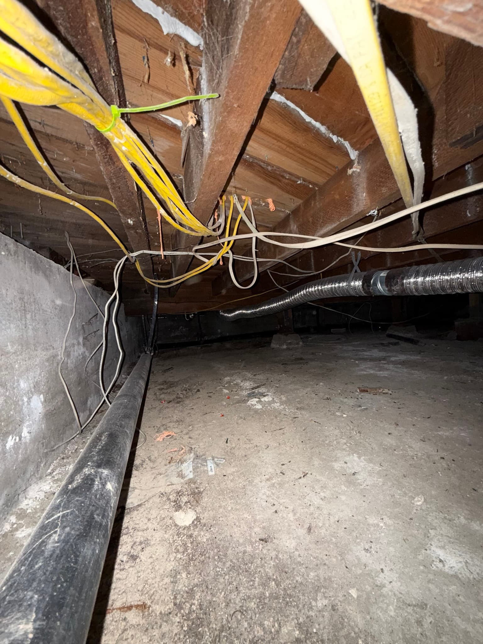 Crawl space or underfloor area showing a flexible metal dryer vent duct running unsupported across wooden floor joists, with multiple yellow and white electrical conductors routed loosely nearby