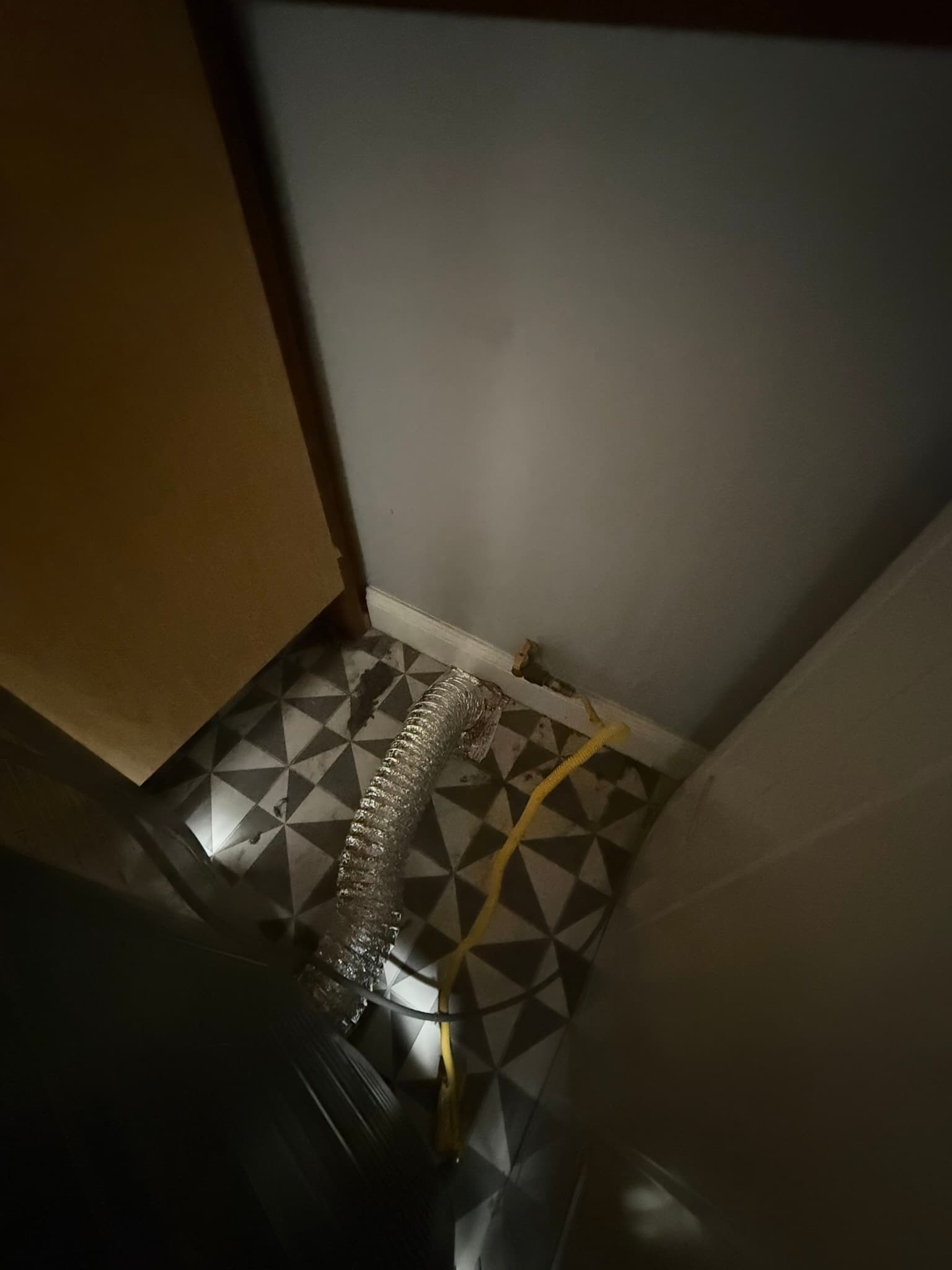 Flexible foil dryer vent duct routed along a basement or crawl space floor near a foundation wall, with a yellow cable visible alongside the ductwork