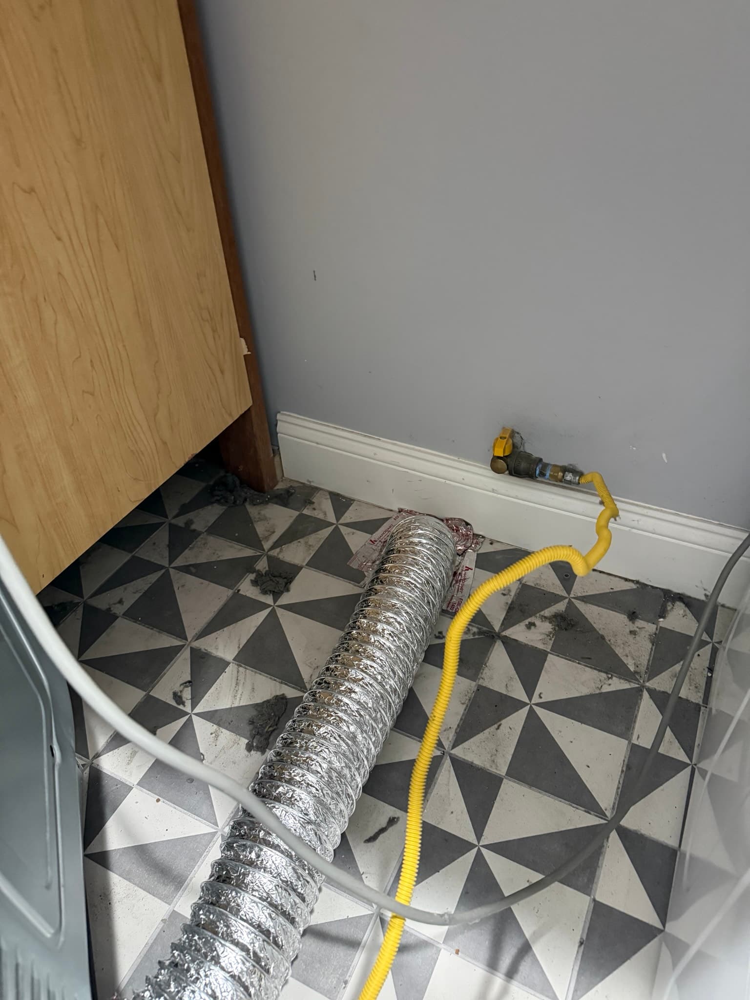 Flexible metal dryer vent duct with heavy lint and dust buildup on its exterior surface, routed across a tiled interior floor with yellow plastic ducting attached at one end