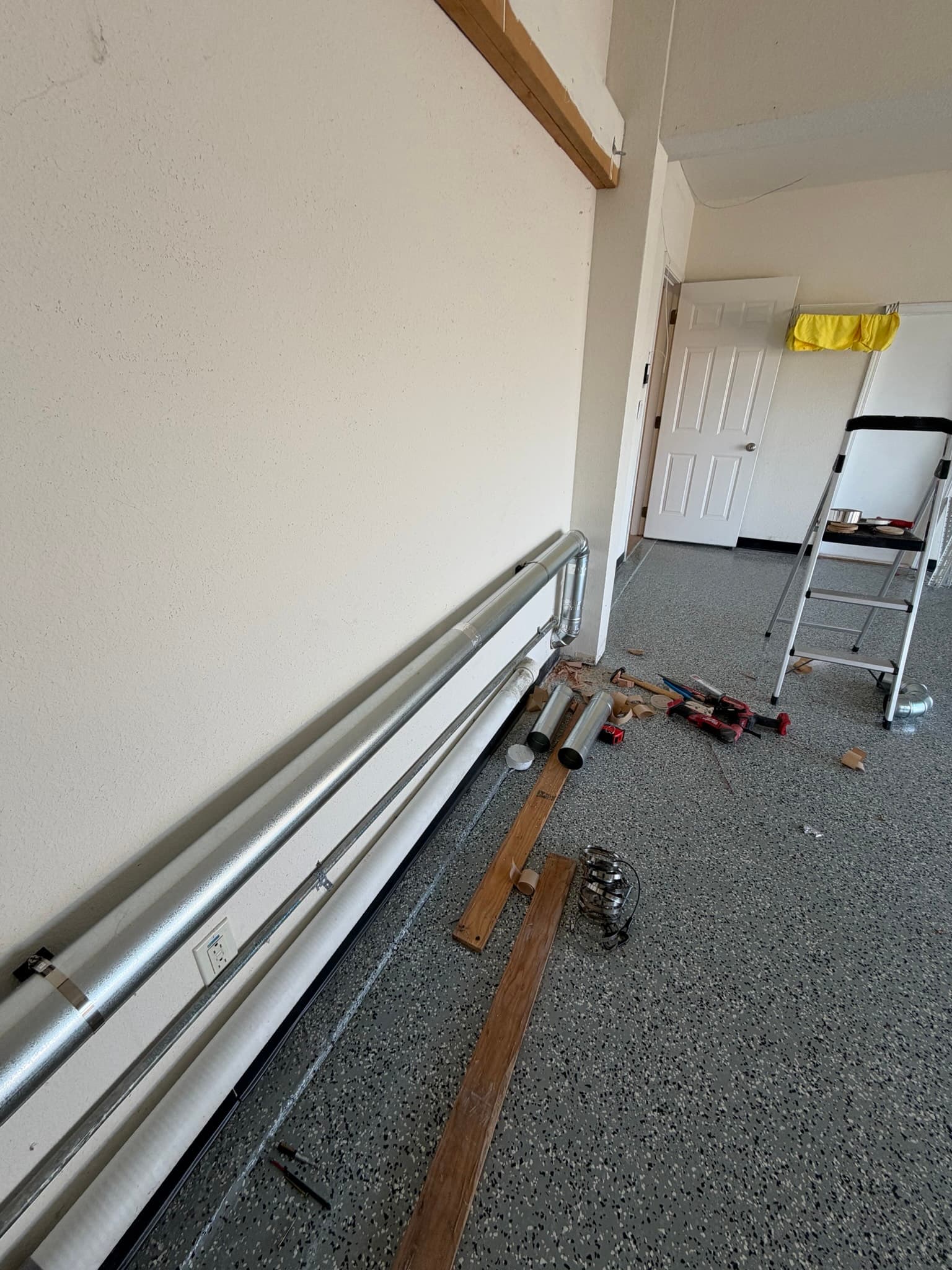 This image shows metal dryer exhaust ducting laid out on a basement floor during an installation or repair project. Two sections of rigid metal duct are visible alongside wooden framing materials and various fasteners and clamps. The ducts appear clean and undamaged, with proper connectors ready for assembly. This appears to be pre-installation staging rather than an in-situ inspection photo.