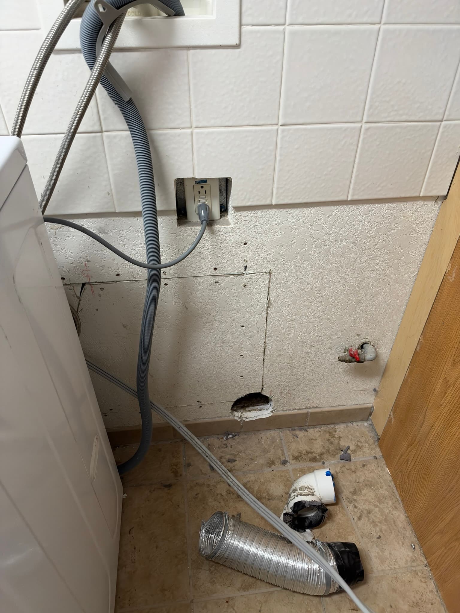 This photo shows a dryer exhaust installation with multiple code violations and maintenance issues. Two flexible metal ducts are visible: one appears disconnected or loosely fitted at the wall penetration (center), and another aluminum flexible duct lies on the floor below. The wall opening shows visible dirt, debris, and discoloration around the duct entry point. The installation lacks a proper exterior termination cap or periscope vent, and the ducts appear unsecured and improperly routed, creating fire hazard and exhaust flow concerns.