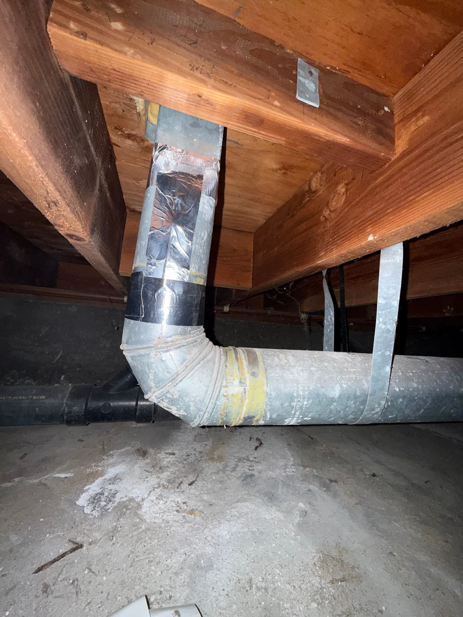 Flexible dryer vent ductwork in a crawl space with a sealed joint wrapped in yellow foil tape, showing heavily dust-coated exterior surfaces and a vertical-to-horizontal duct transition