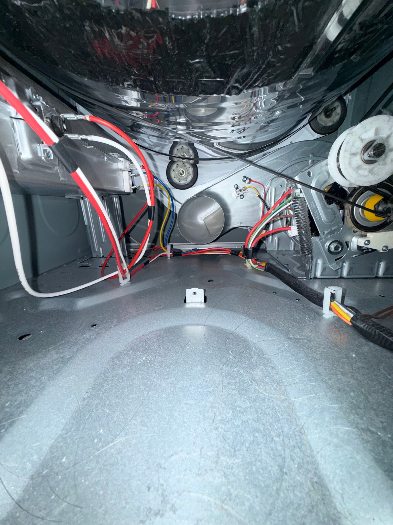 Interior view of a dryer cabinet from below, showing the drum, felt seals, pulleys, and color-coded wiring with some lint accumulation on interior surfaces