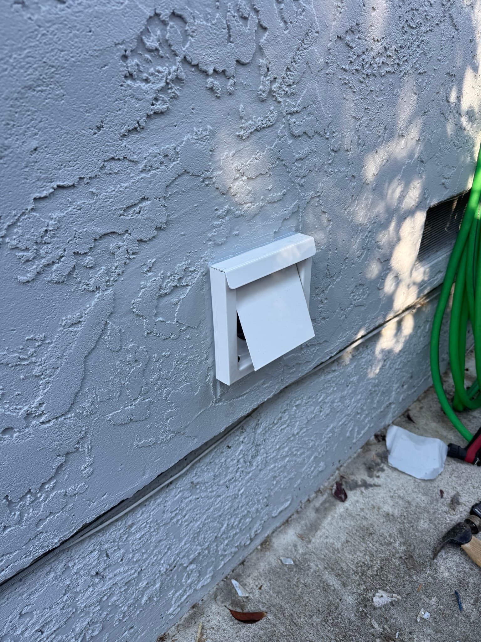White Innovate dryer wall vent cap on a gray stucco wall at ground level with the dryer running, damper flap open and allowing exhaust airflow to escape freely to the exterior