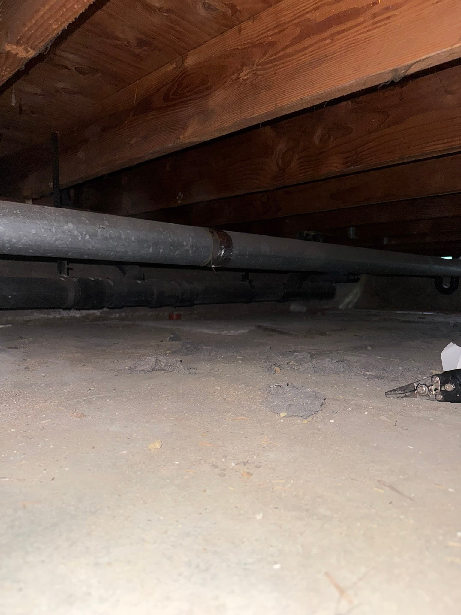 Metal ductwork resting near a dusty concrete floor in a crawl space or basement, with darkened wooden floor joists above and visible surface oxidation on the duct exterior