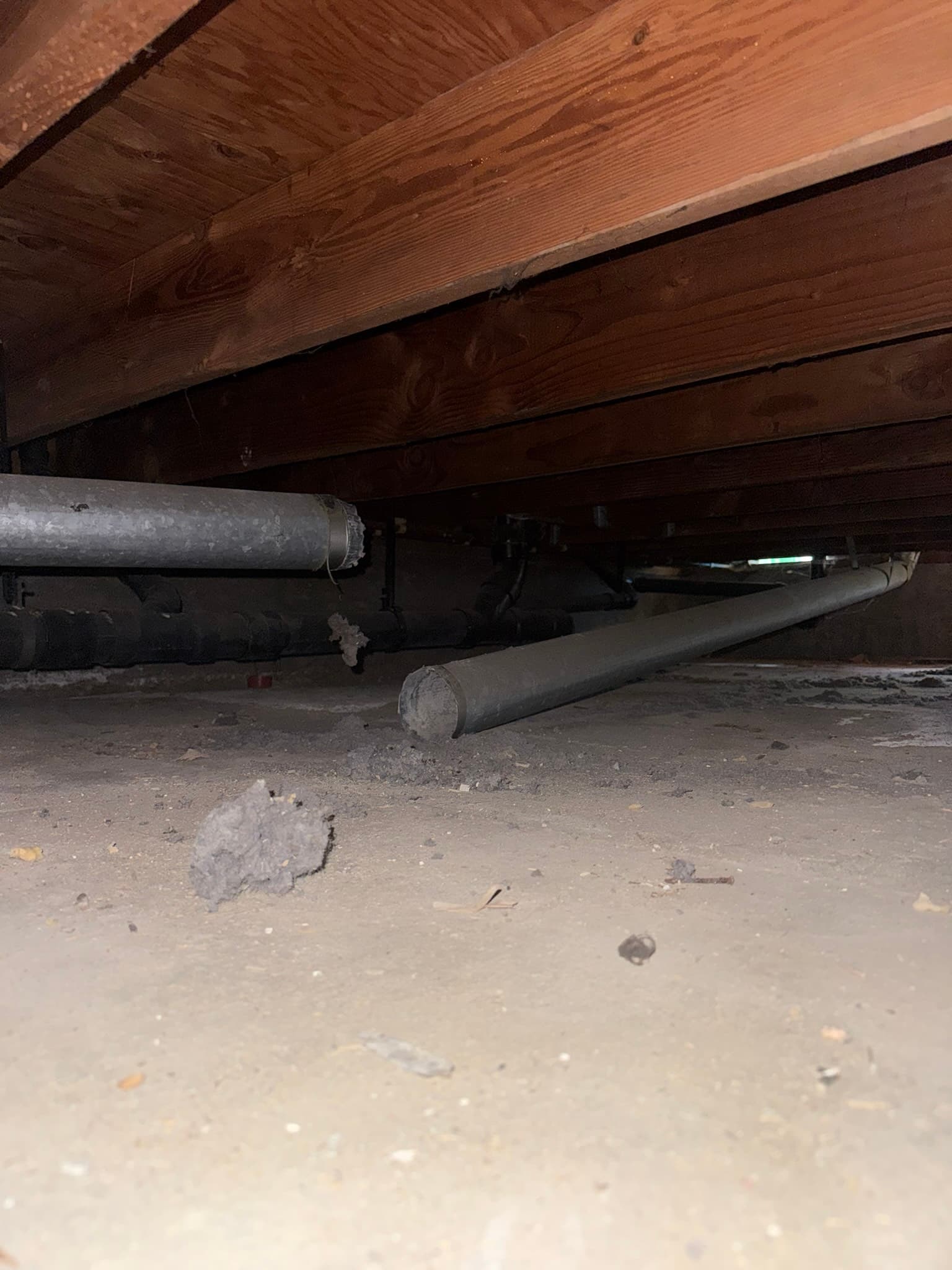 Rigid metal ductwork in a crawl space beneath wooden floor joists, with visible surface corrosion, dust accumulation, and loose insulation debris on the floor below