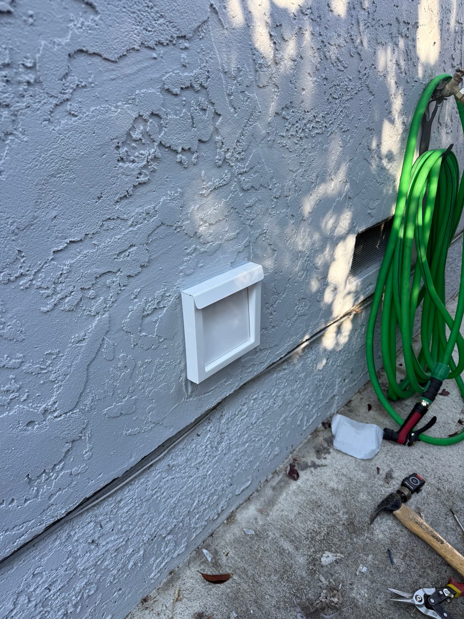 White exterior dryer vent cap installed on a textured stucco wall at standard height above grade, with a coiled green garden hose and tools on the ground below indicating an active service call