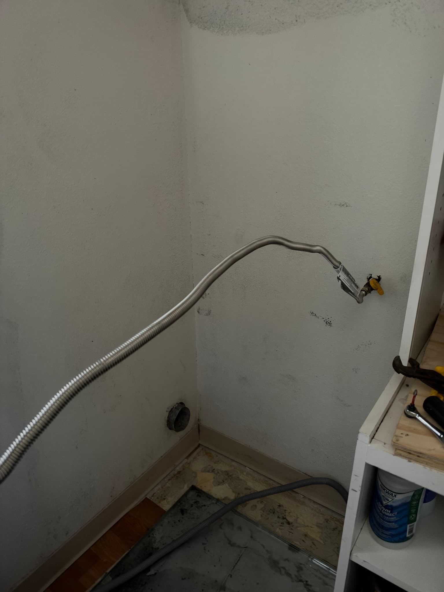 Newly installed 8 foot stainless steel washing machine supply line running in a corrugated metal hose from the wall shutoff valve down to the laundry standpipe, with a capped dryer exhaust outlet visible in the wall nearby
