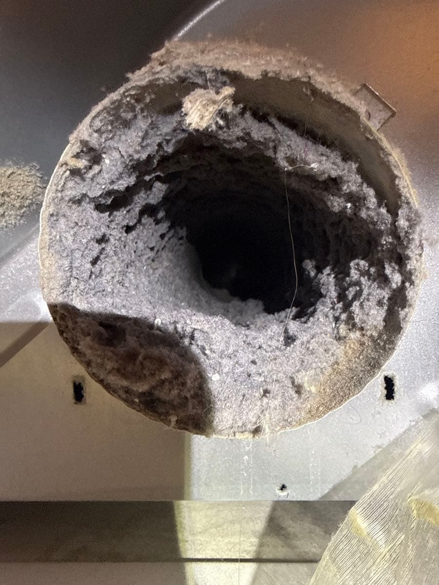 Photo of a dryer vent wall opening heavily clogged with compacted lint debris coating the perimeter and duct interior, with a thin wire probe inserted into the duct opening