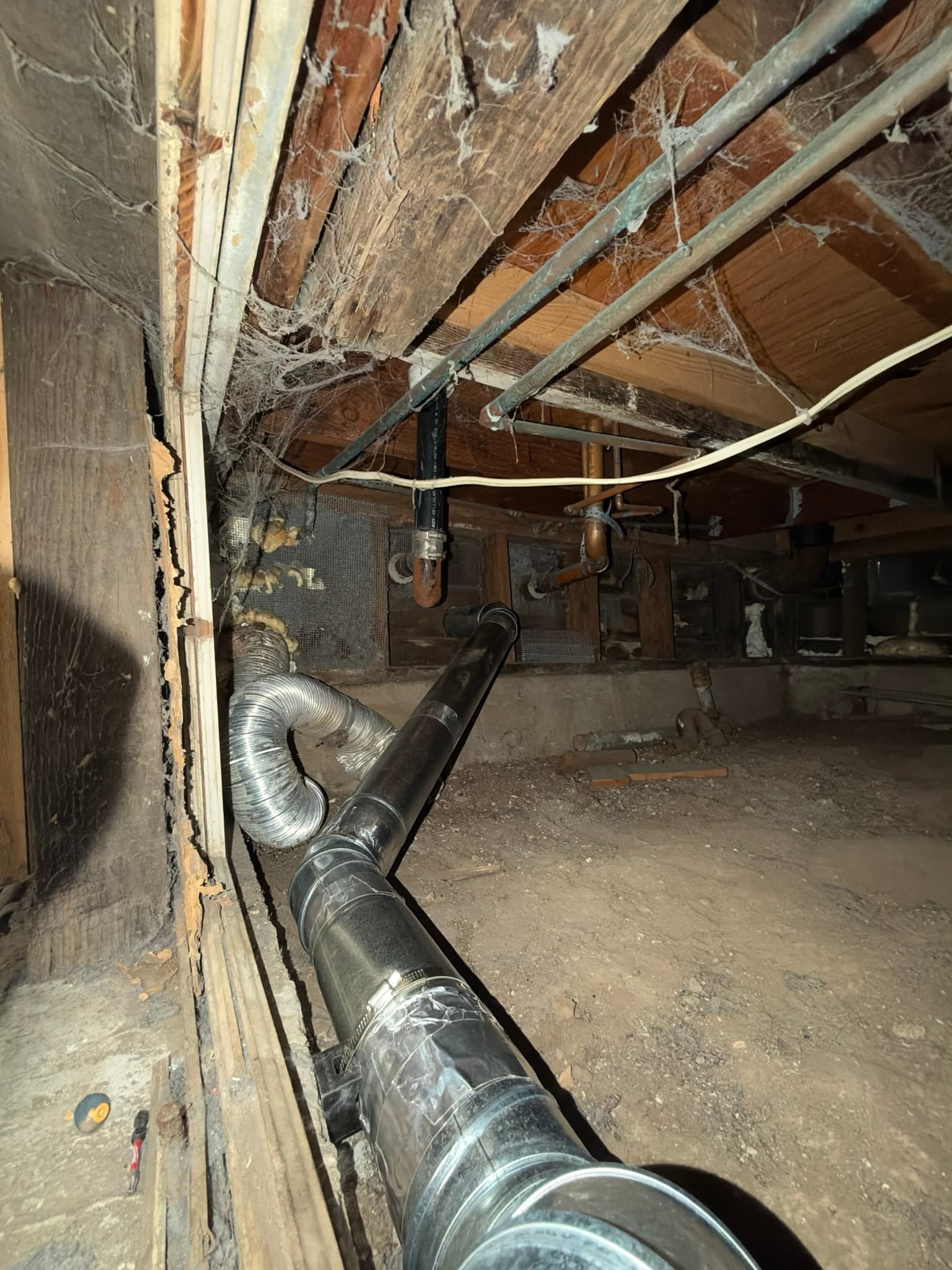 Crawlspace view of newly installed rigid galvanized snap lock duct connected to a short run of flexible transition duct, sloped toward the exterior wall termination.