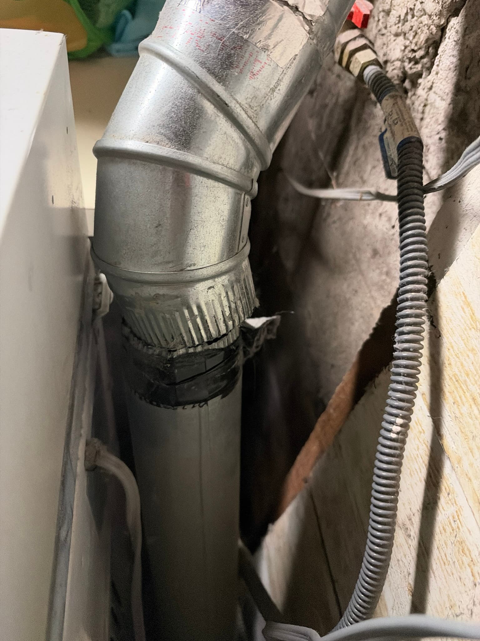 Rigid metal elbow fitting connecting to ductwork behind the dryer, with the joint wrapped in tape near exposed wall insulation and wiring.