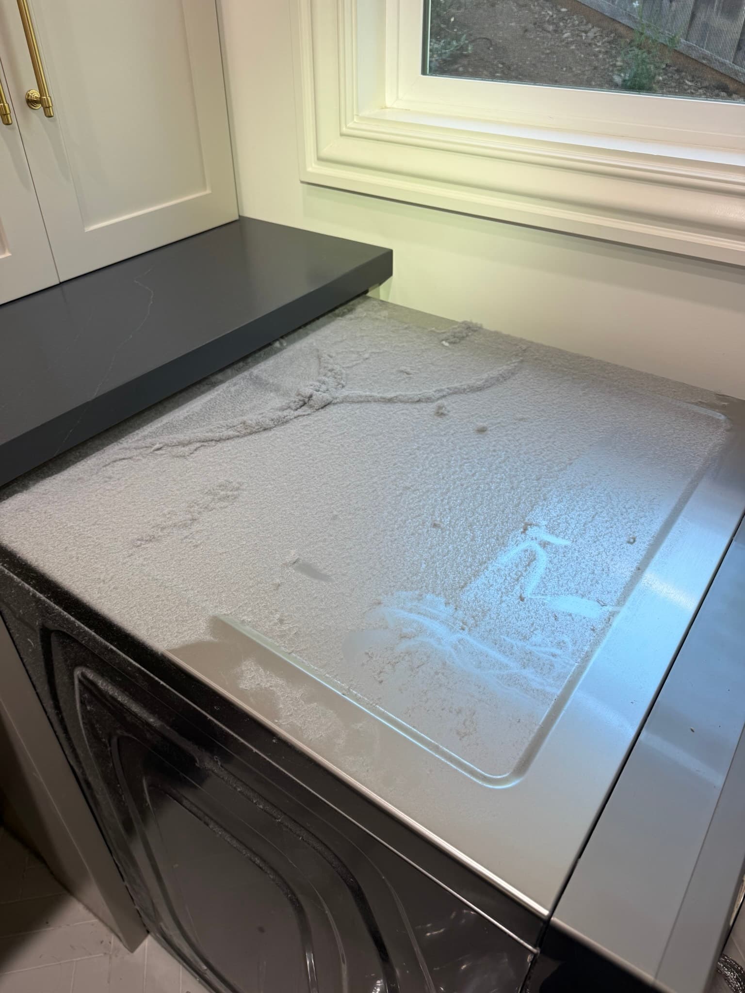 Top surface of a clothes dryer coated with a heavy layer of gray lint and dust, with the lint trap area visibly clogged, indicating long-term inadequate dryer vent maintenance and an elevated fire risk
