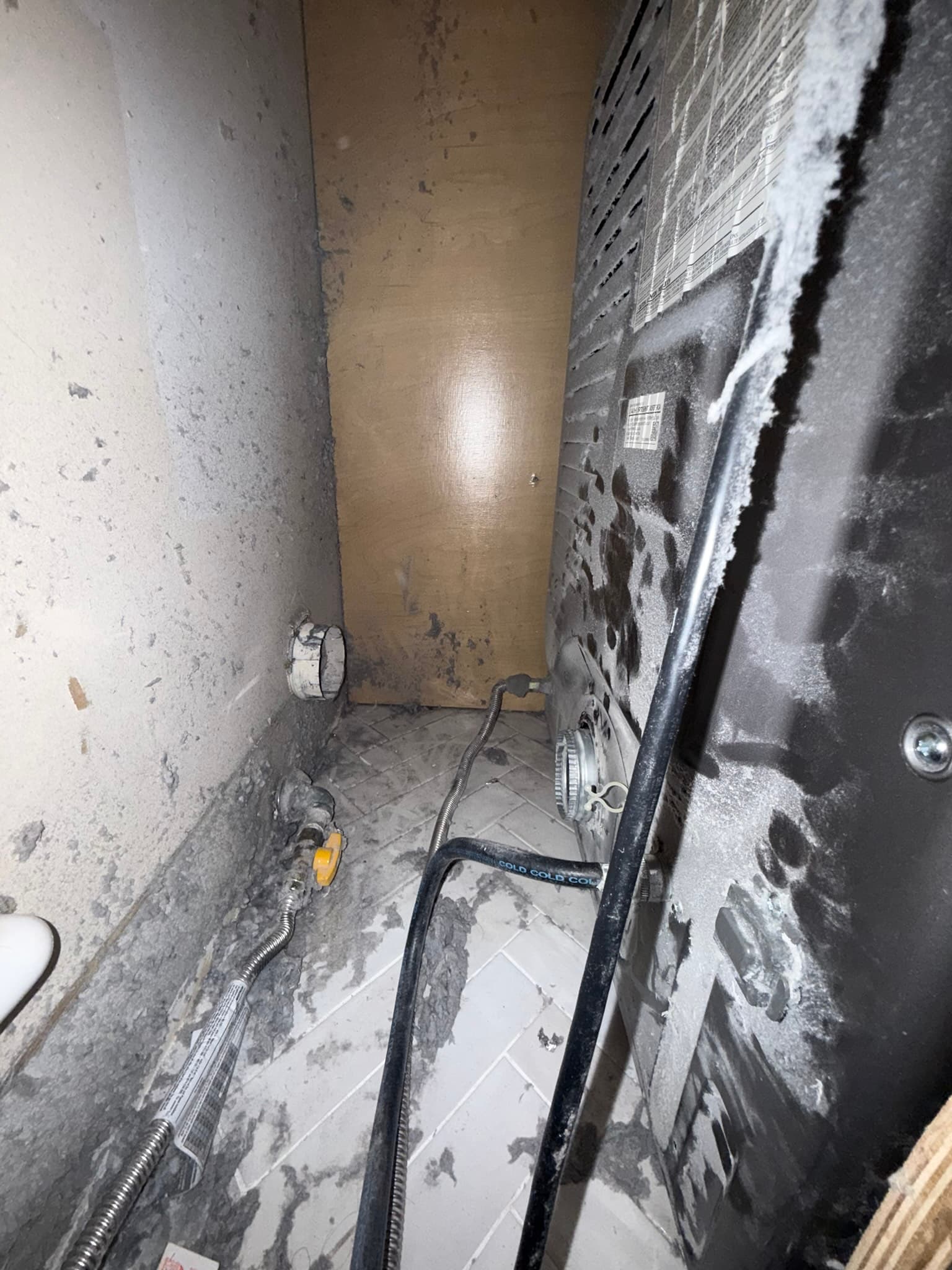 Narrow utility space behind a dryer showing heavy lint and dust accumulation on the floor and walls, with a black flexible metal exhaust duct running along the floor and a separate flexible hose visible to the left, indicating a long-term disconnected duct condition