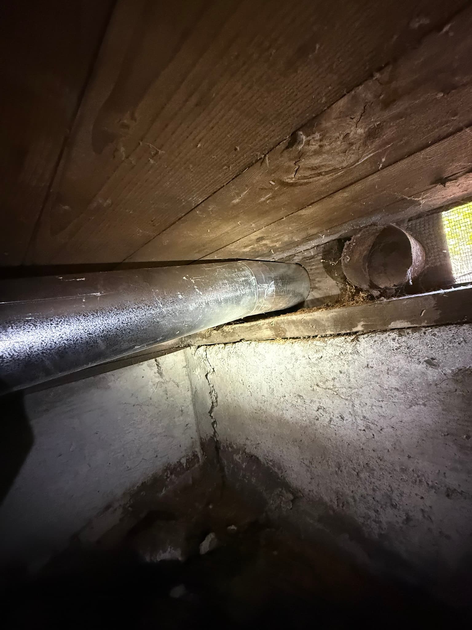 Flexible metallic dryer exhaust duct in a crawl space connecting to a corroded rigid metal duct termination, with rust and debris accumulation visible around the opening and dust on wooden framing above