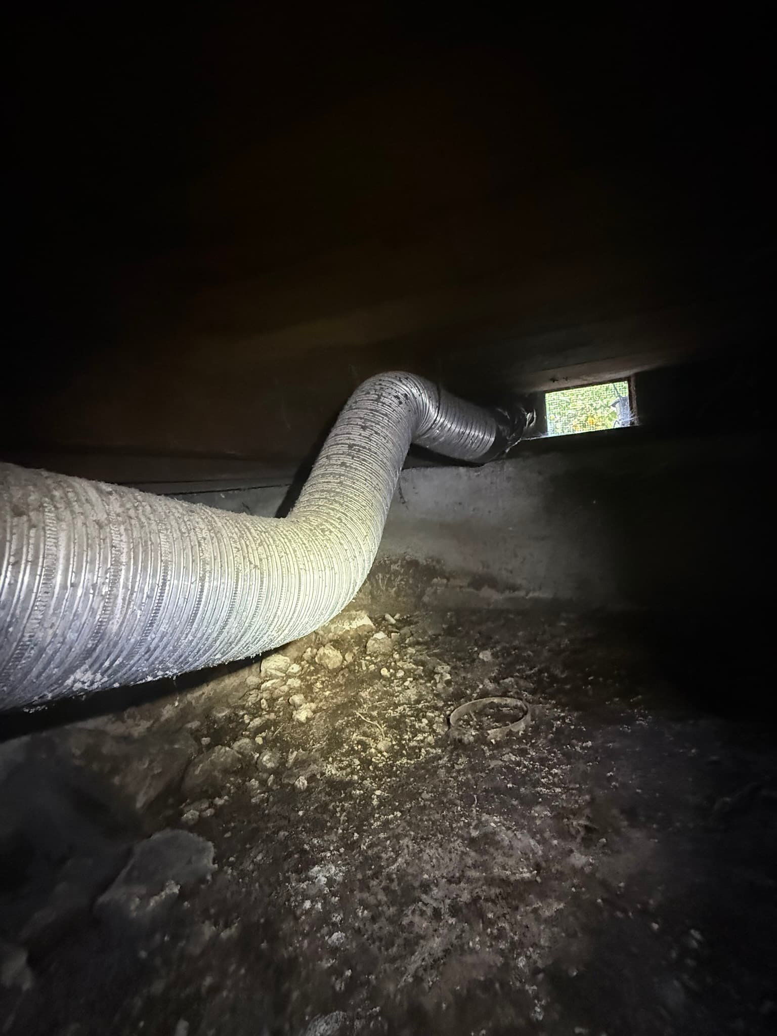 Old semi-rigid dryer duct in a crawl space, visibly rusted, bent out of alignment, and separated from the wall vent opening, with lint debris on surrounding surfaces