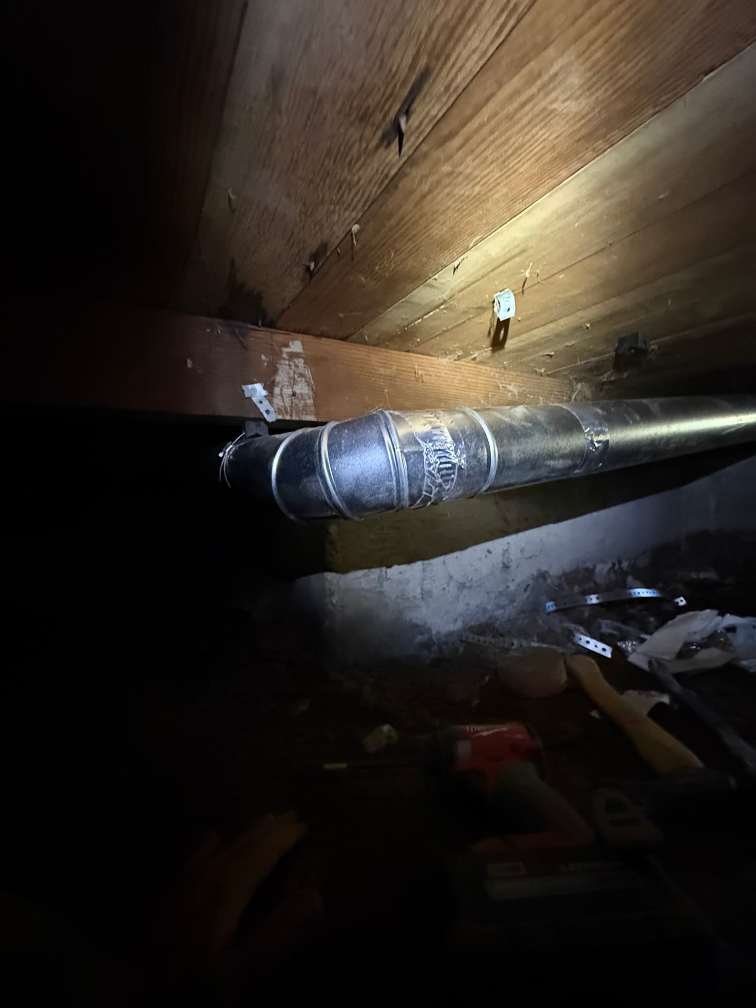 Flexible dryer duct running horizontally through a dark crawl space, secured with a clamp connector, showing dust and debris accumulation on its exterior surface