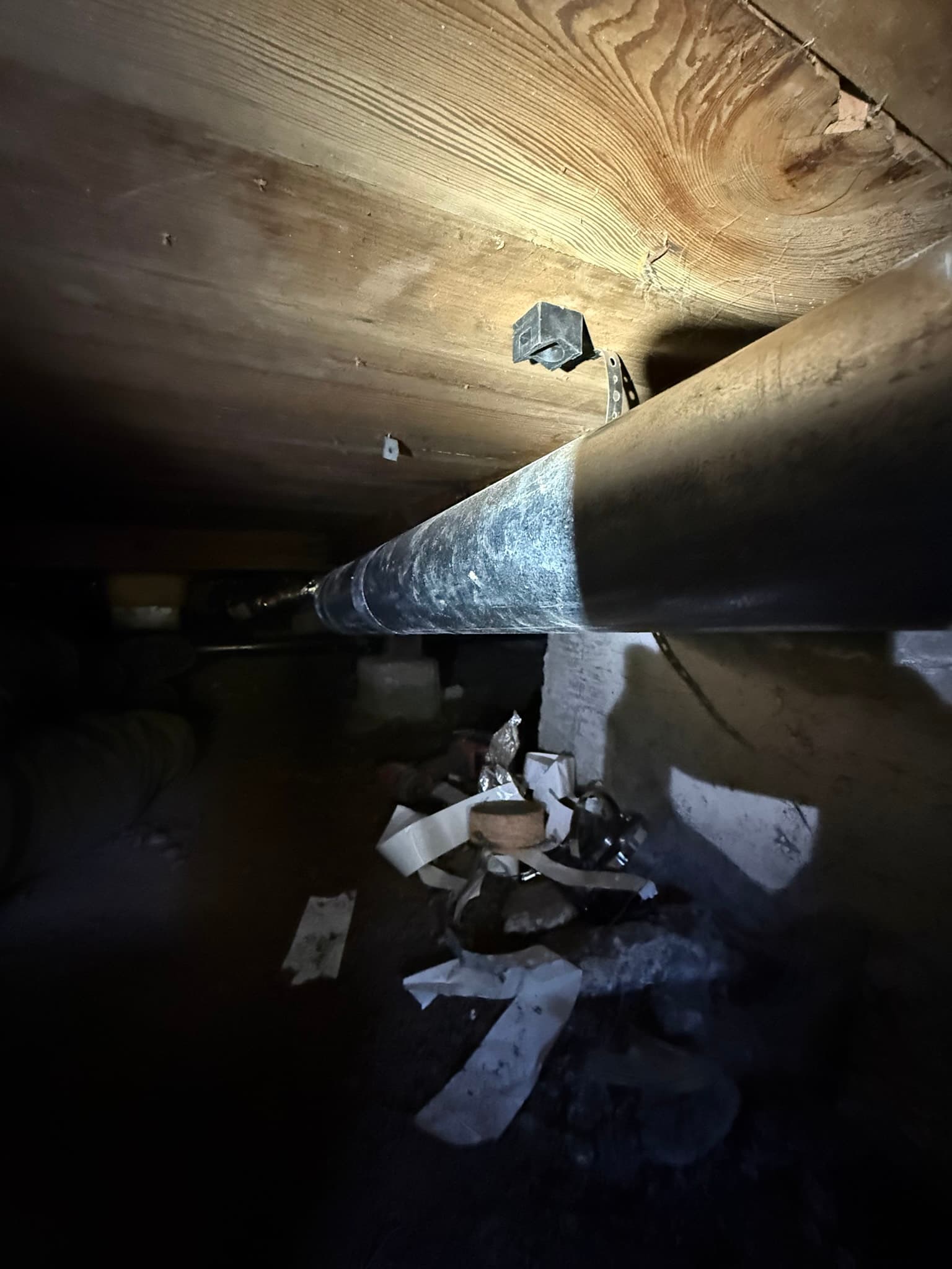 Flexible metal dryer duct in a crawl space connecting to a metal vent box mounted to the wooden ceiling structure, with lint and loose debris visible on surrounding surfaces and gaps at connection points