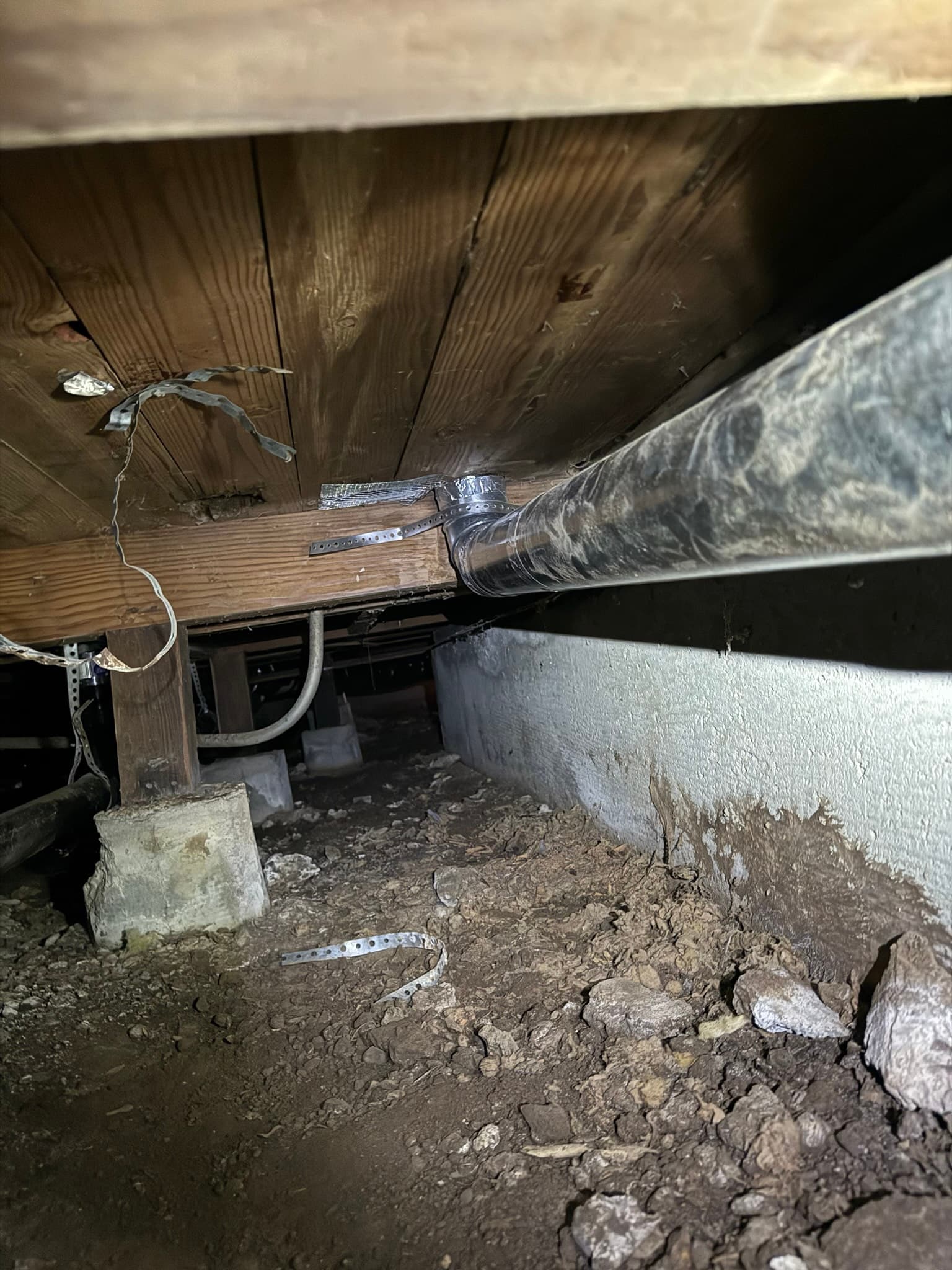 Crawl space floor penetration showing heavy lint accumulation around a disconnected flexible dryer duct with exposed wooden framing and loose strapping visible nearby