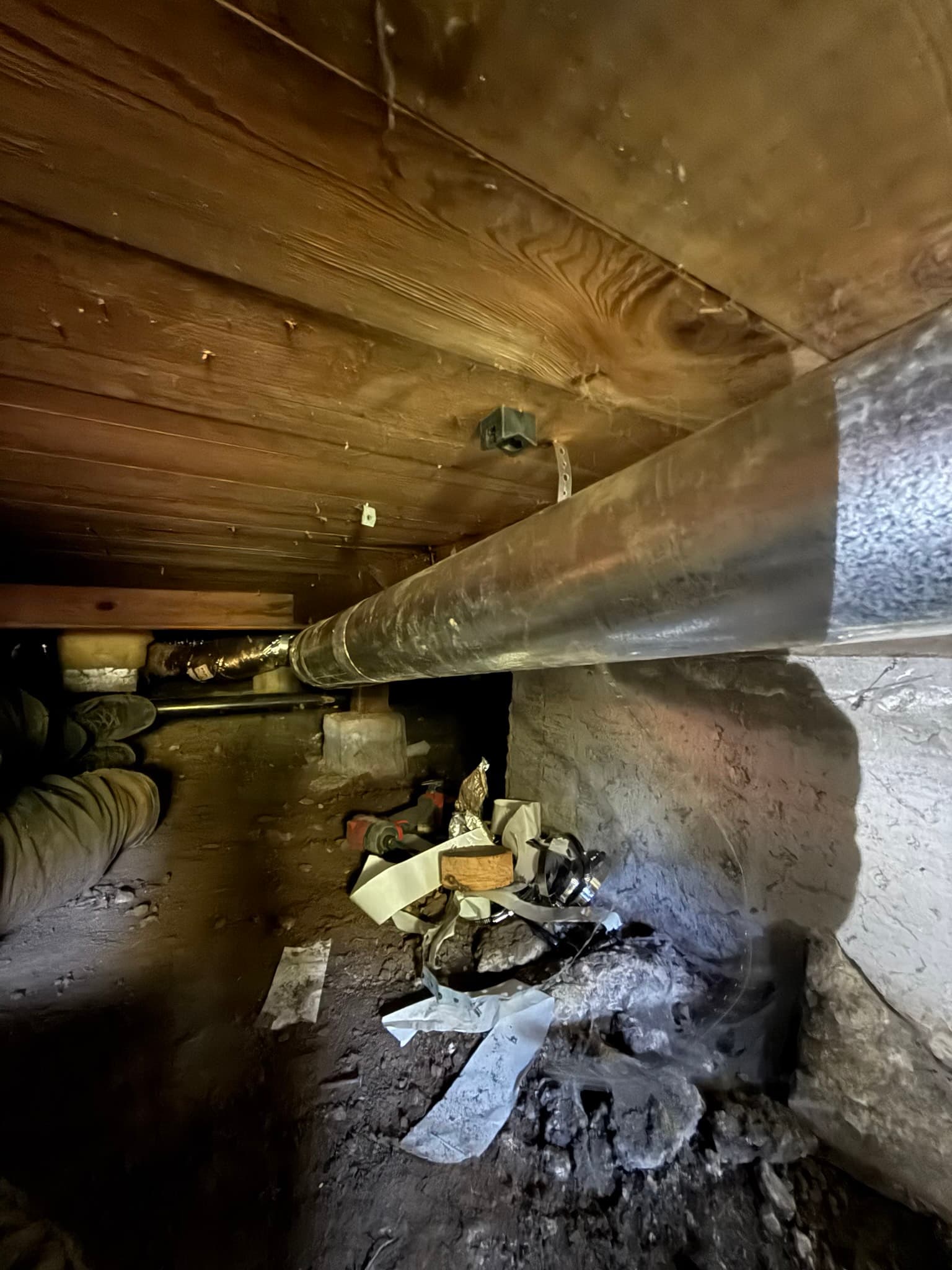 Heavily lint-coated flexible dryer duct running horizontally through a dirty crawl space, with debris covering the floor and dust coating the wooden ceiling framing above