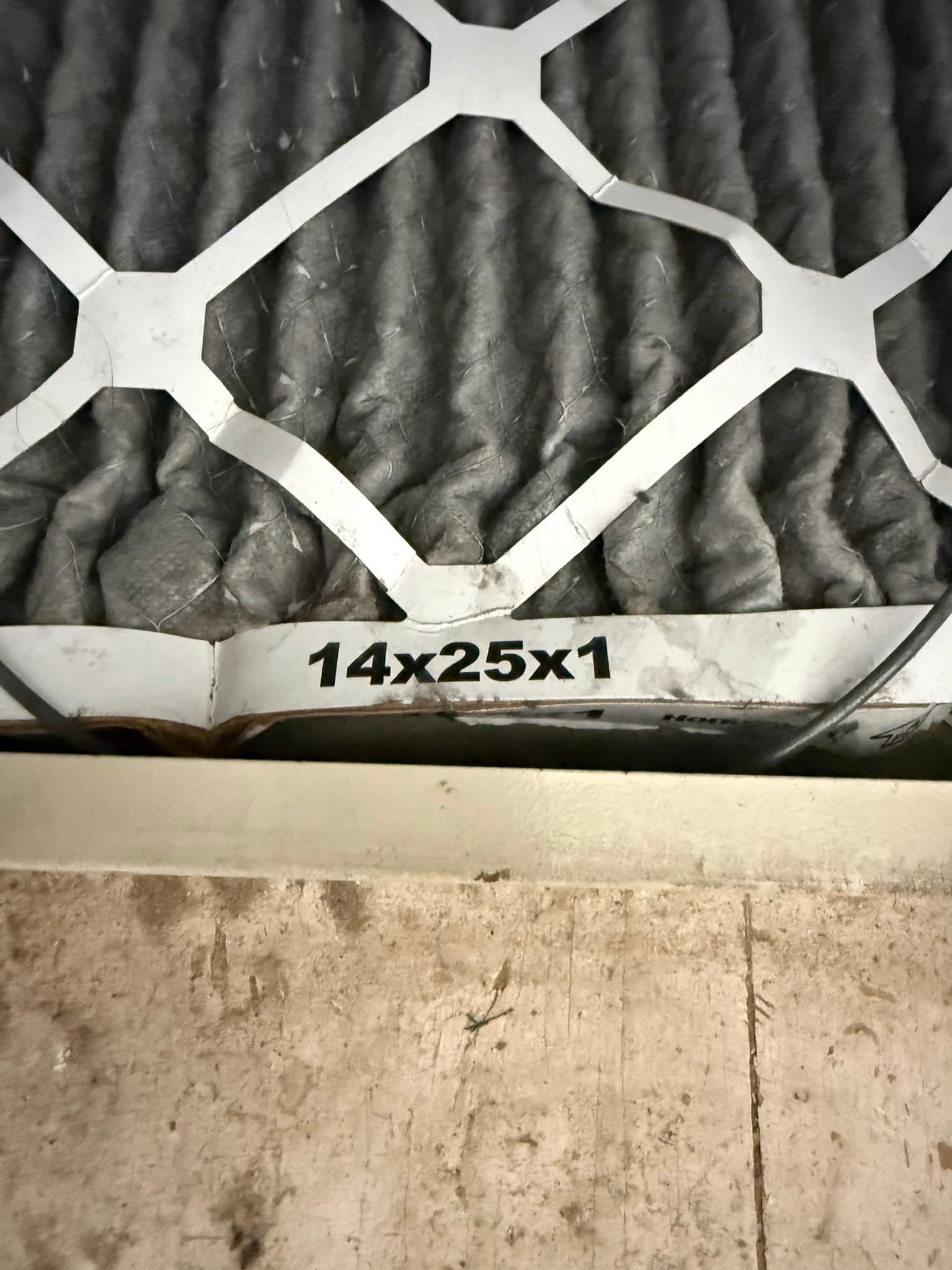 A 14x25x1 residential return-air filter installed against a concrete floor with visible heavy lint and dust accumulation across the pleated surface, indicating the filter has not been replaced in an extended period