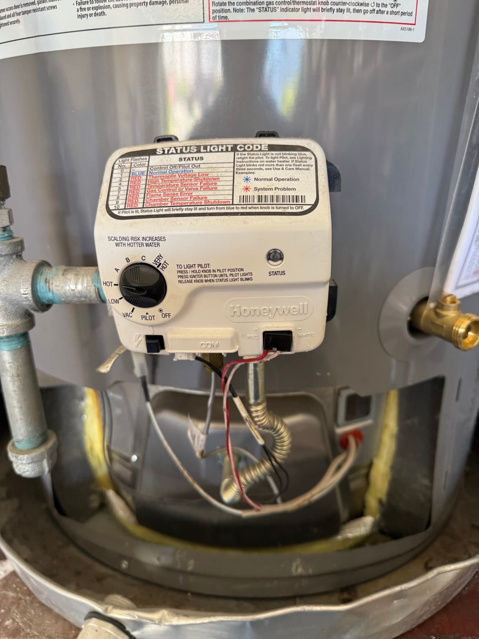 Top of a natural gas water heater showing a Honeywell thermostat control with a rotary temperature dial and status light code chart, with corroded or mineral-scaled metal pipe connections on the left and brass fittings on the right