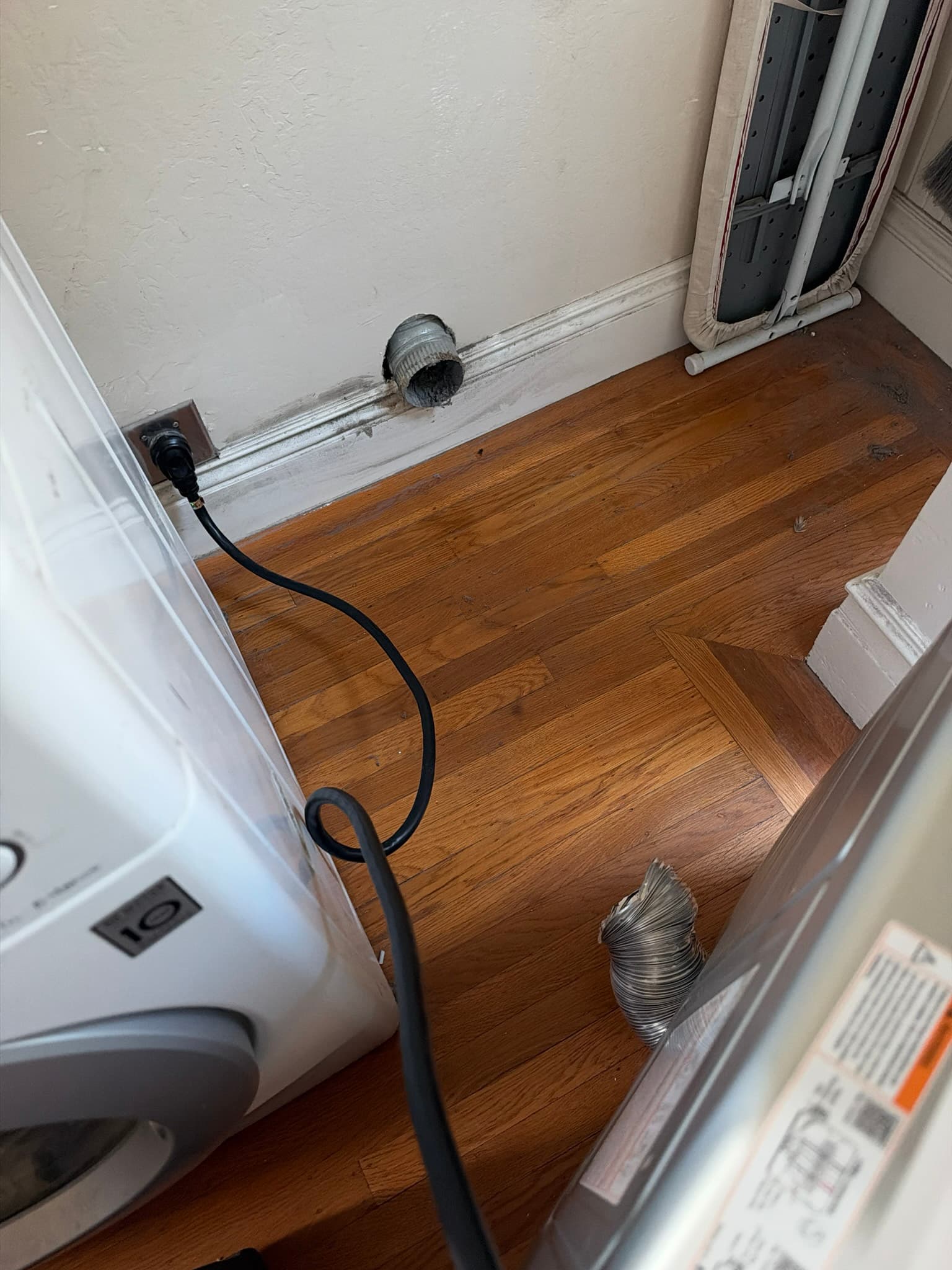 Wall-mounted metal duct collar disconnected from the dryer with visible lint residue around its opening, flexible foil transition duct detached and resting on the floor.