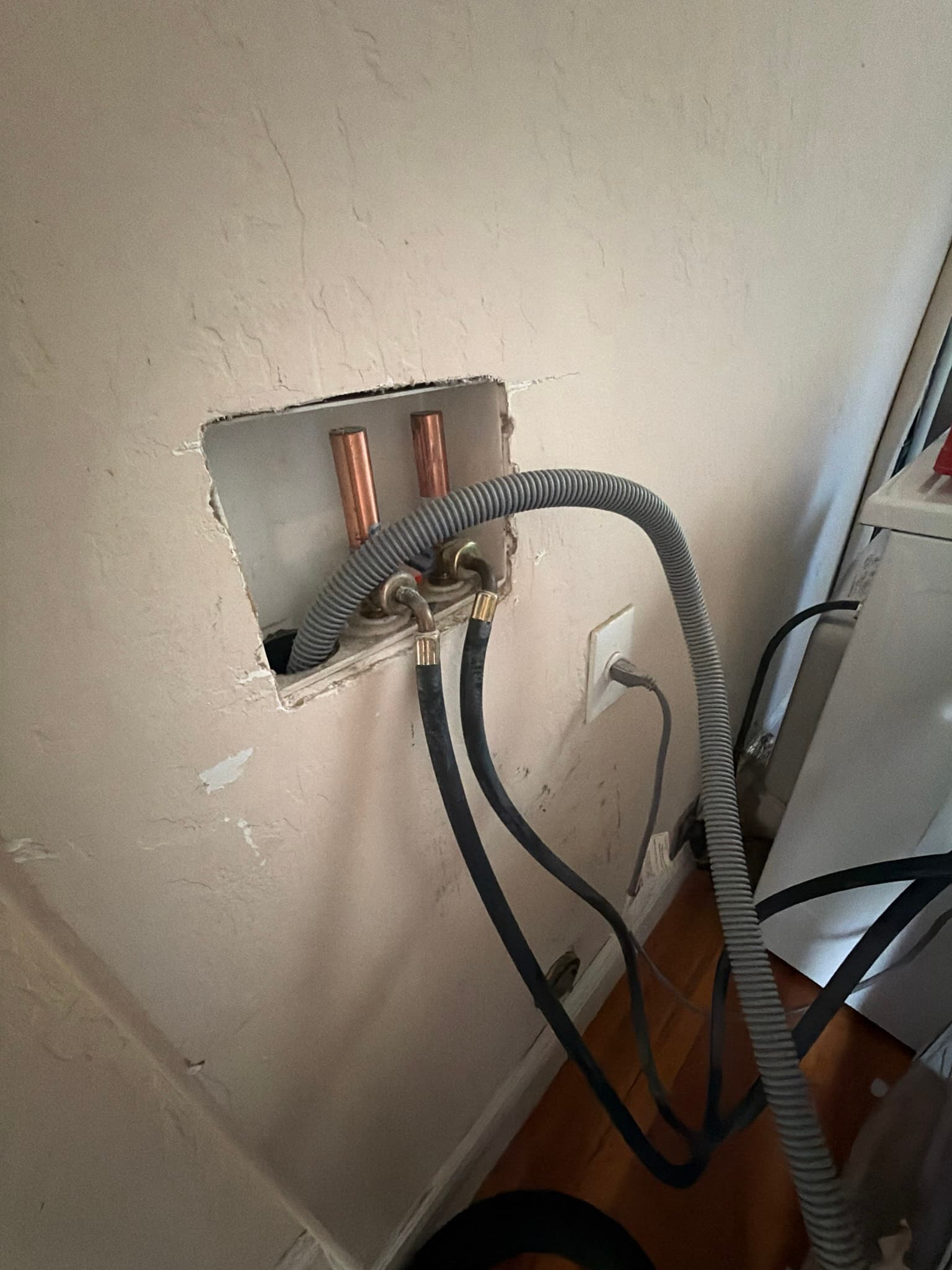 Washing machine supply box with copper stub-outs, hot and cold hoses, and a corrugated drain line routed through an open wall cutout before service.