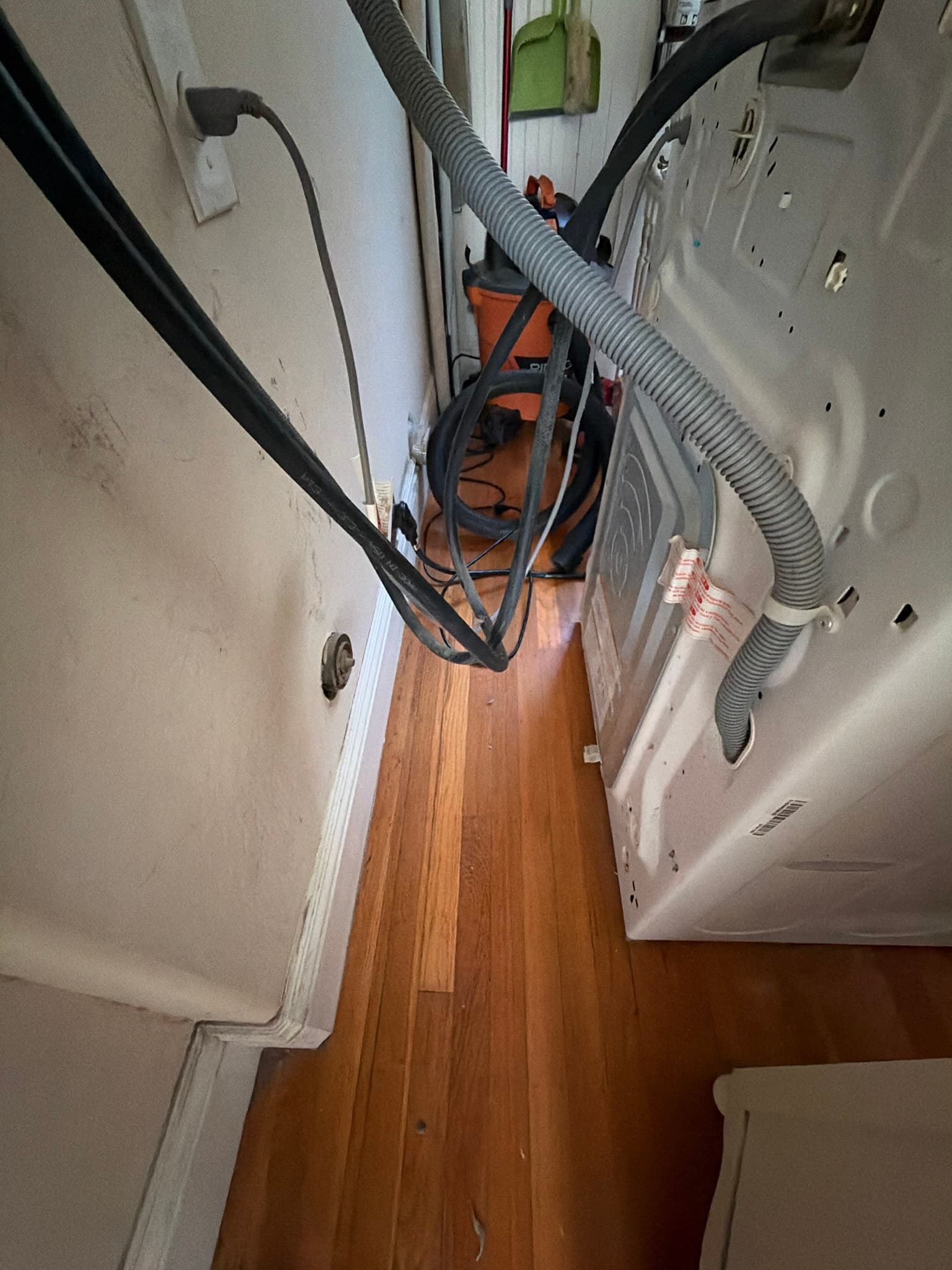 Dryer pulled away from the wall exposing the rear panel and exhaust connection, with a shop vacuum and rotary brush cable staged for cleaning the duct line.