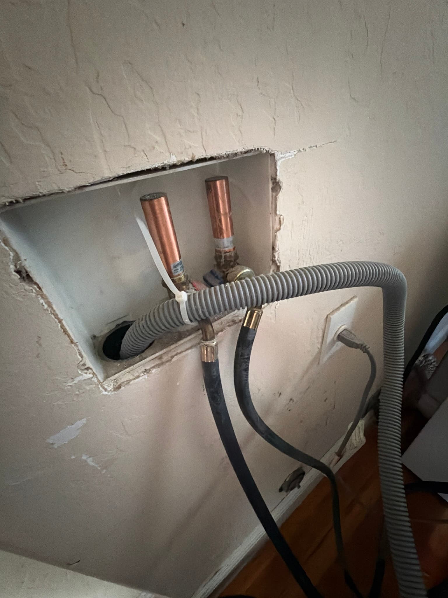 Washing machine supply alcove showing copper water lines, braided supply hoses, an electrical outlet, and a corrugated drain hose secured with a zip tie to keep it seated in the standpipe.