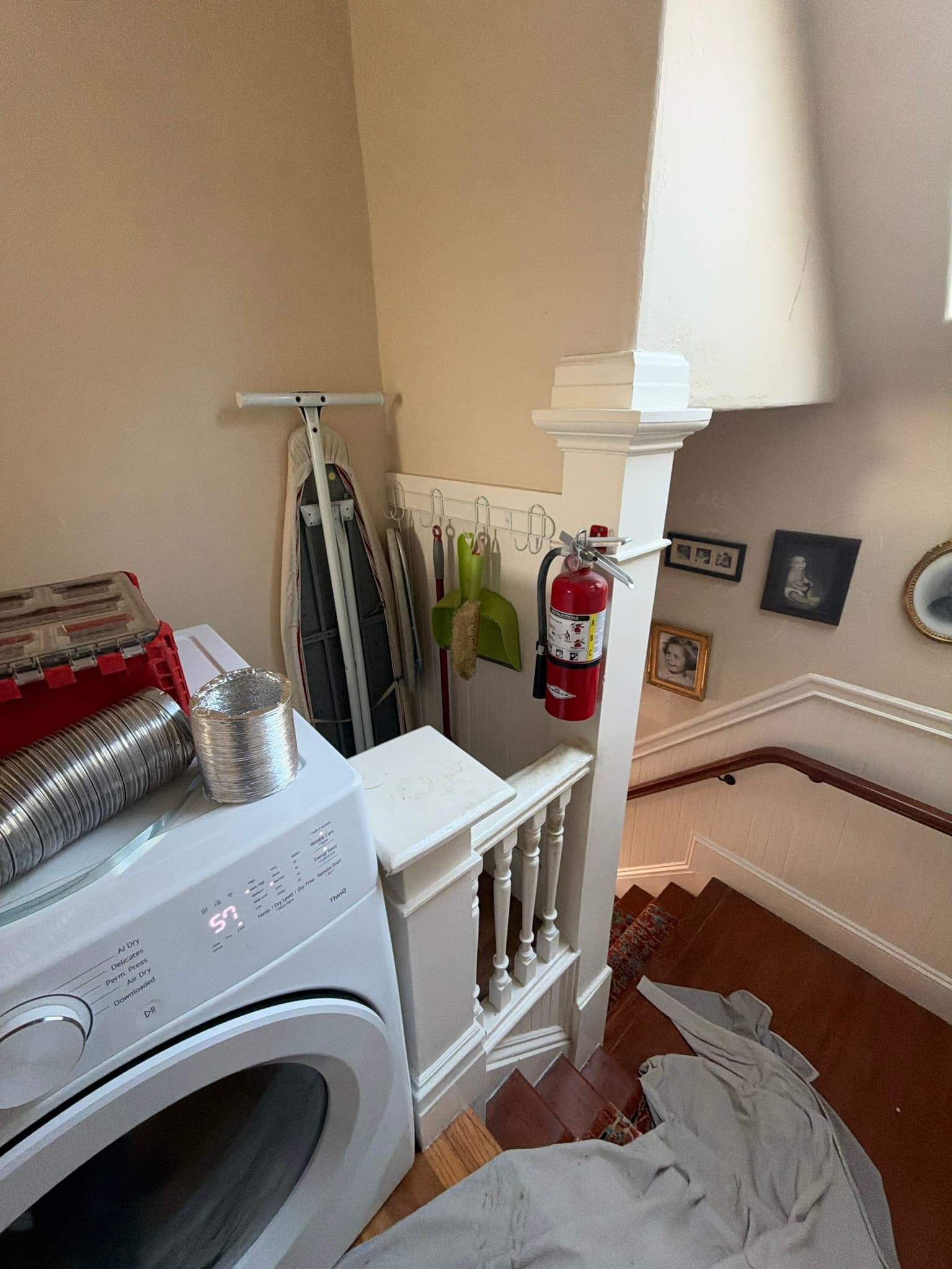 Front-load dryer near a stairway landing with sections of flexible foil transition duct and semi-rigid metal ducting staged on top of the unit during service, stairwell protected with drop cloths.