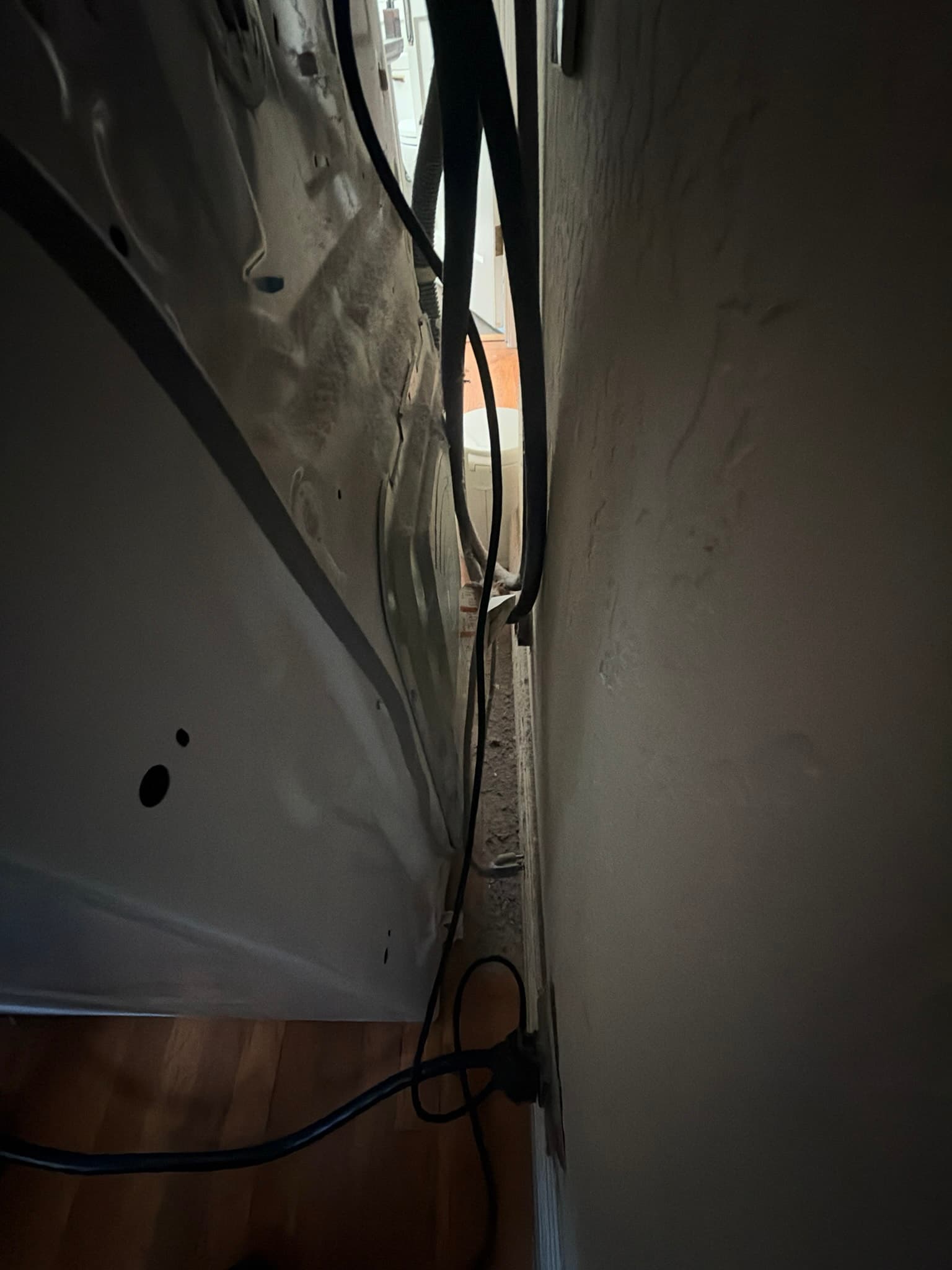 Narrow gap behind the dryer with the unit pulled away from the wall, showing the rear panel, power cord, and dust along the floor and baseboard.