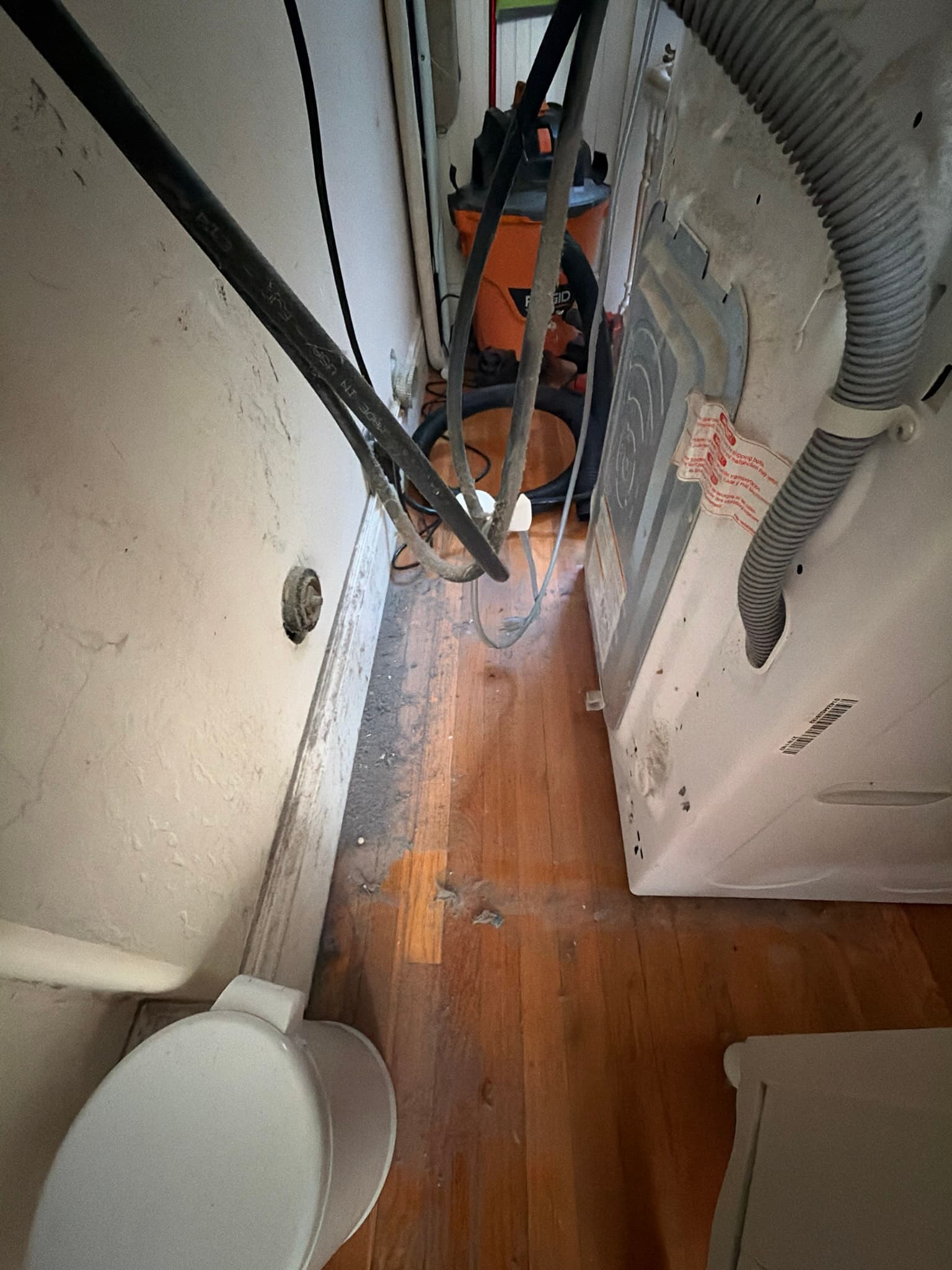 Before photo of the dryer pulled from the wall showing the transition duct connection and a wall opening coated with built-up lint and dust along the baseboard, with a shop vacuum staged for cleaning.