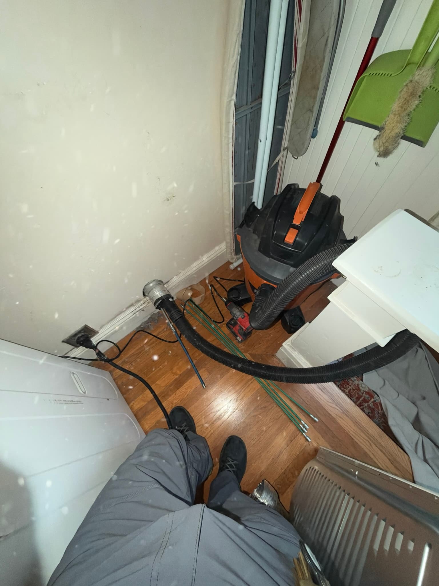 Mid-service photo of active dryer vent cleaning with a shop vacuum hose and drill-powered rotary brush fed through the wall duct opening, lint scattered on the floor nearby.