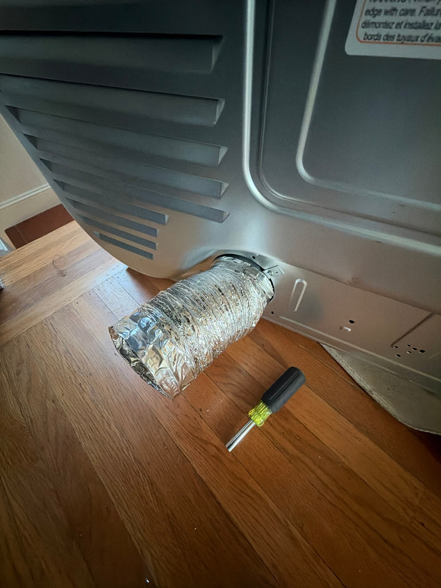 Dryer pulled away from the wall with the flexible foil transition duct disconnected from the rear exhaust port and laid on the hardwood floor for inspection, screwdriver nearby.