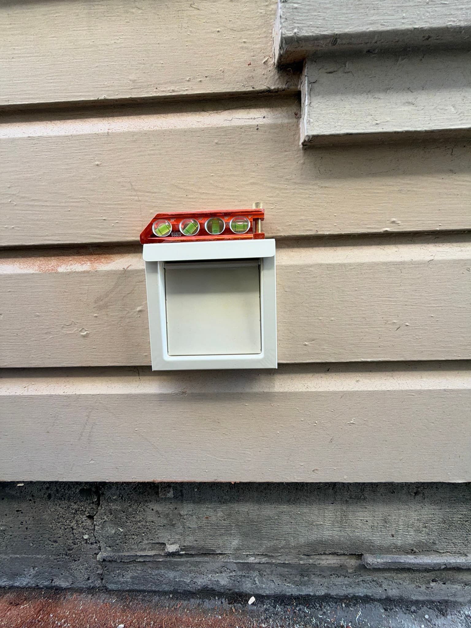 New white flush mount 4 inch dryer wall vent installed on exterior siding with a level resting on top confirming a square, properly seated installation