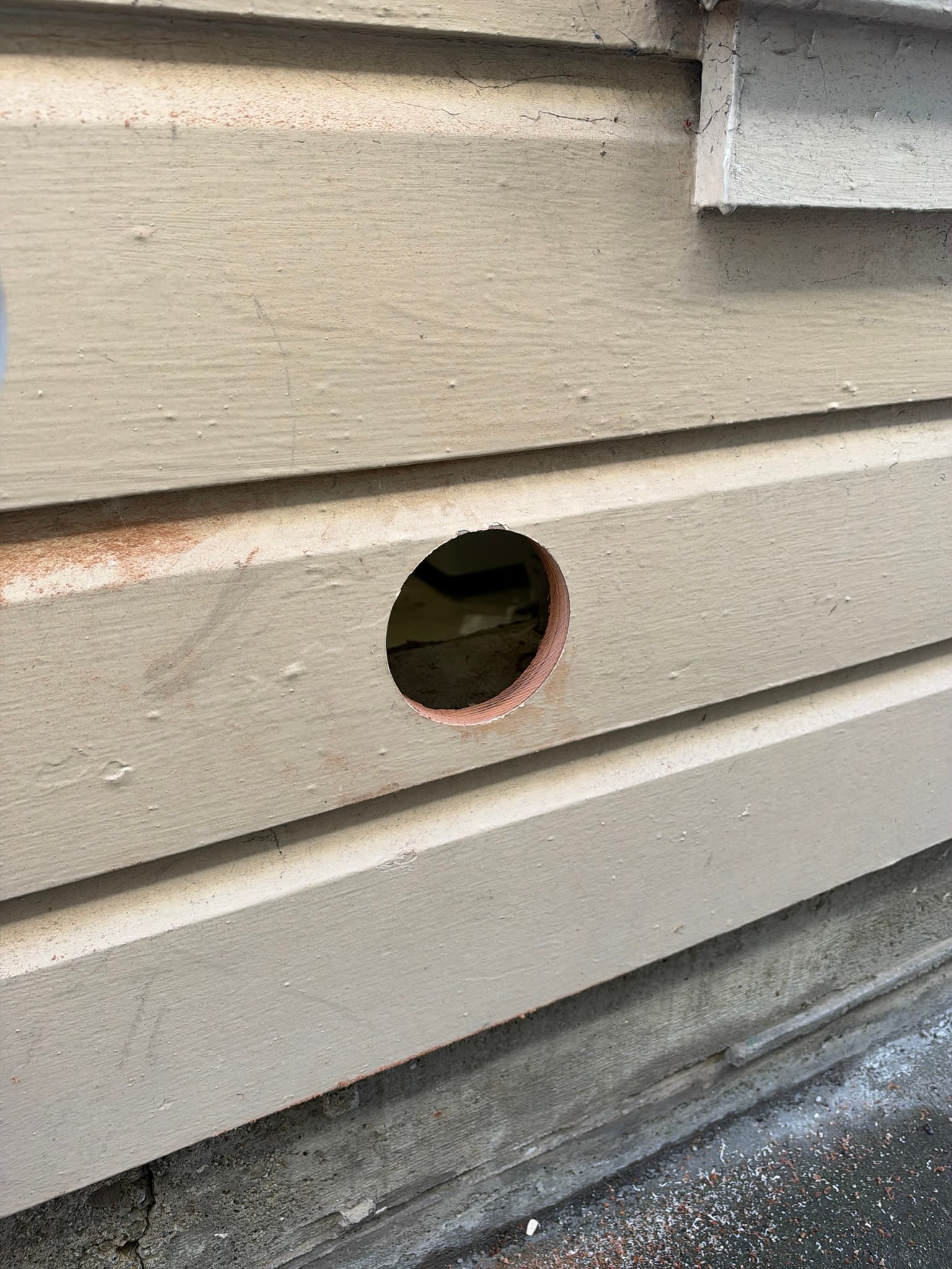 Exterior wall penetration with no vent cap installed, showing a bare hole through the siding with rust staining and the duct visible inside