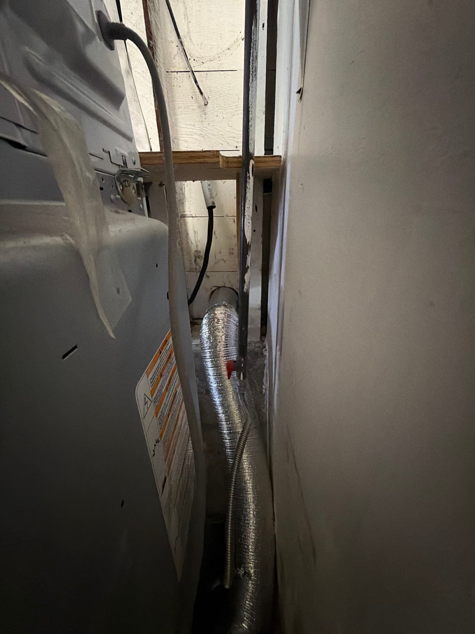 Dryer pulled away from the wall showing flexible foil transition duct routed through a tight cavity to the new wall vent termination