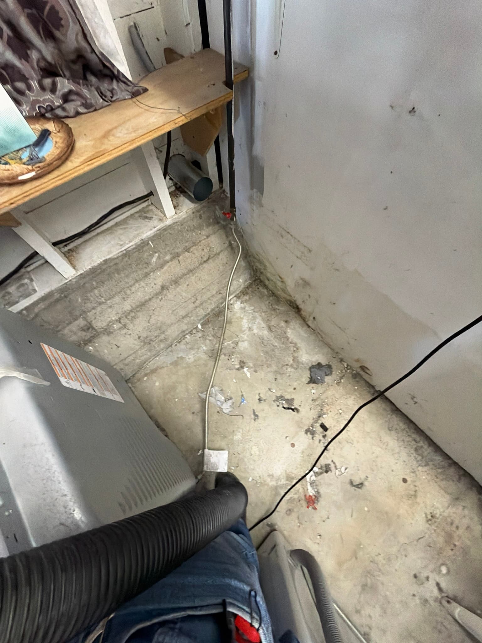 Area behind the dryer before cleaning, showing scattered lint debris on the floor and a flexible transition duct running to the exterior wall opening