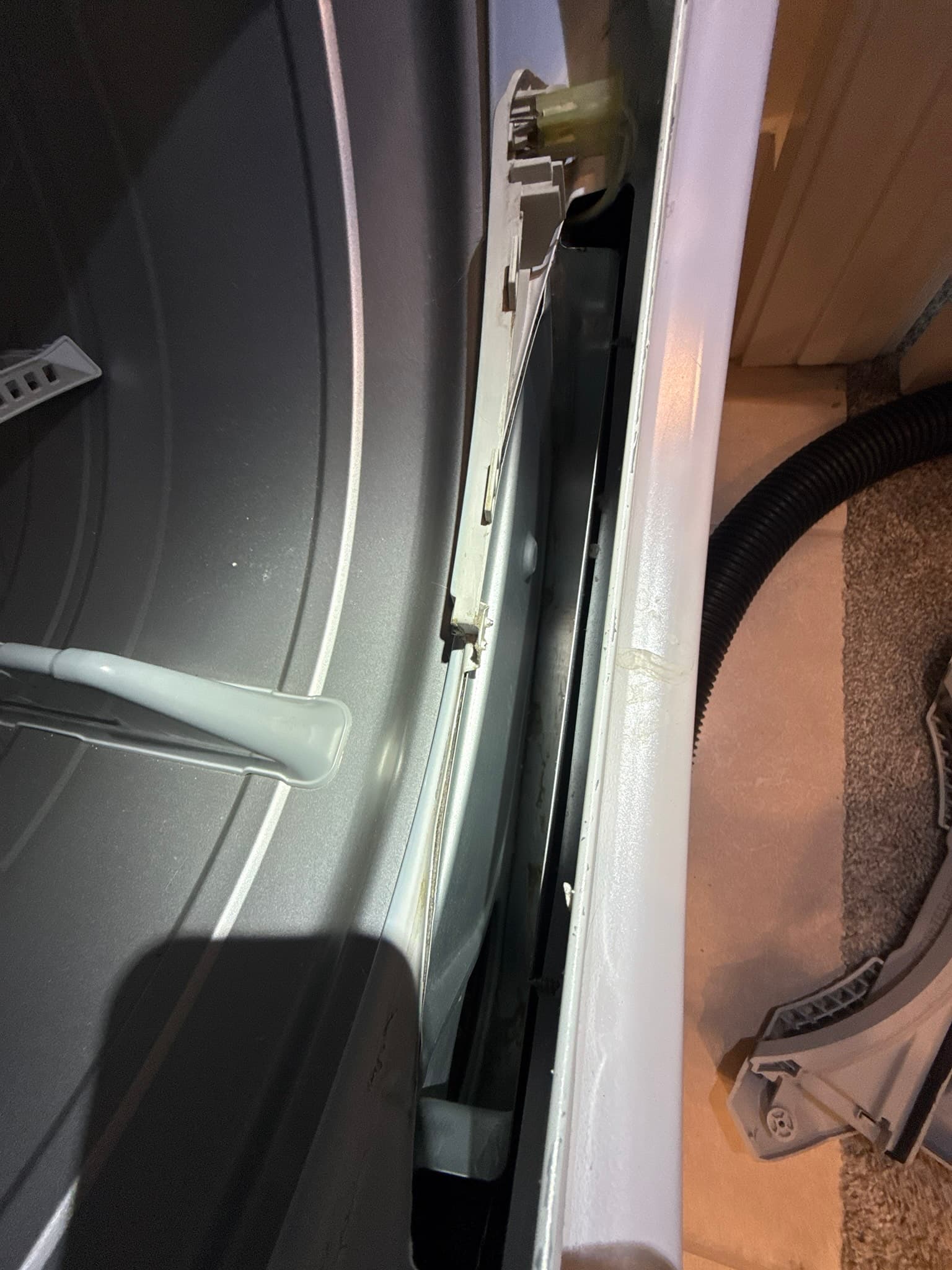 Rear of a residential dryer showing a black flexible corrugated exhaust duct extending from the dryer outlet and connecting to a metal periscope-style vent hood mounted through the wall