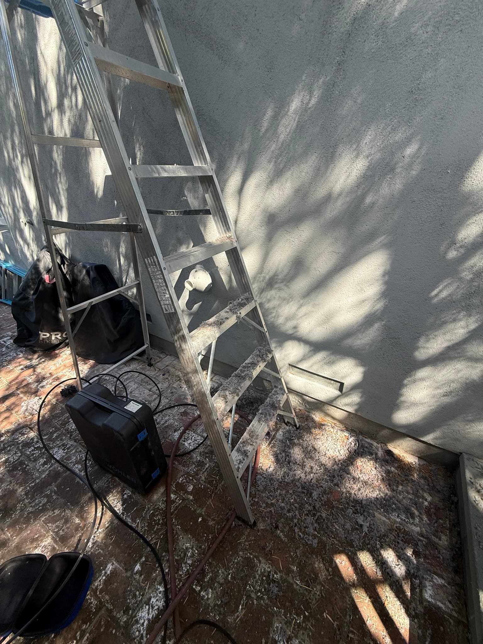 Exterior wall access point showing a rectangular duct opening with a metal trim collar, surrounded by weathered concrete foundation, with a ladder and equipment visible nearby during the cleaning service