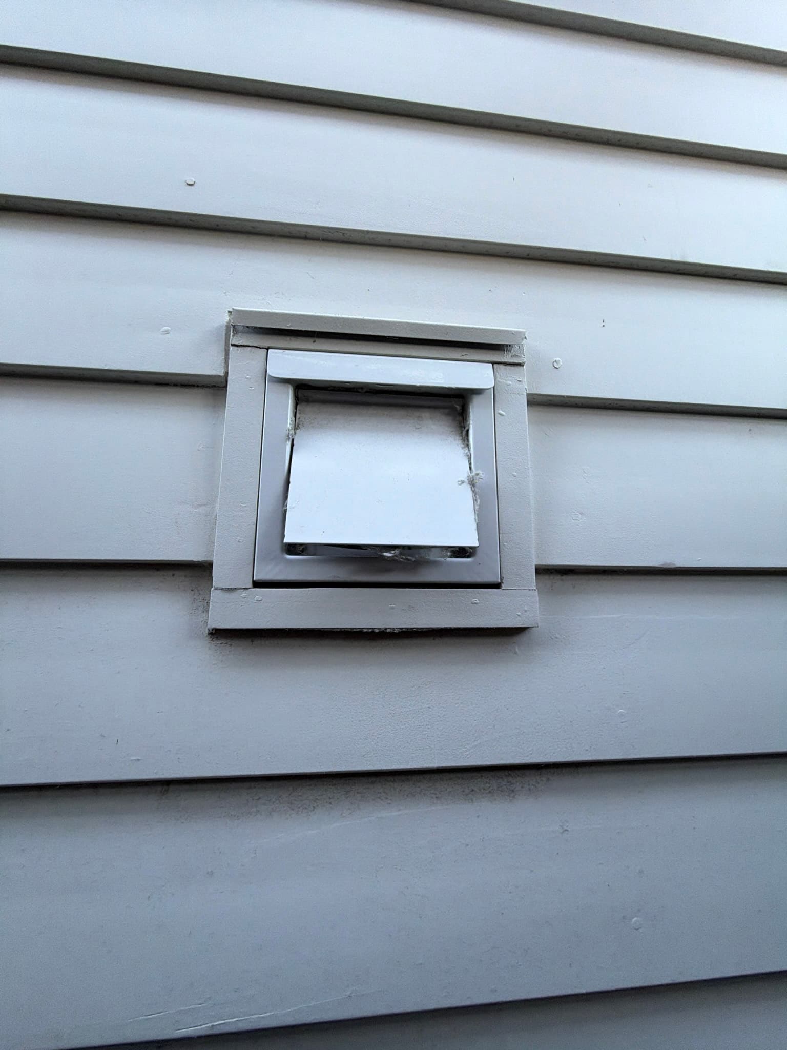 White flush-mounted dryer wall vent in vinyl siding with the damper flap held open by trapped lint and debris at the hinge