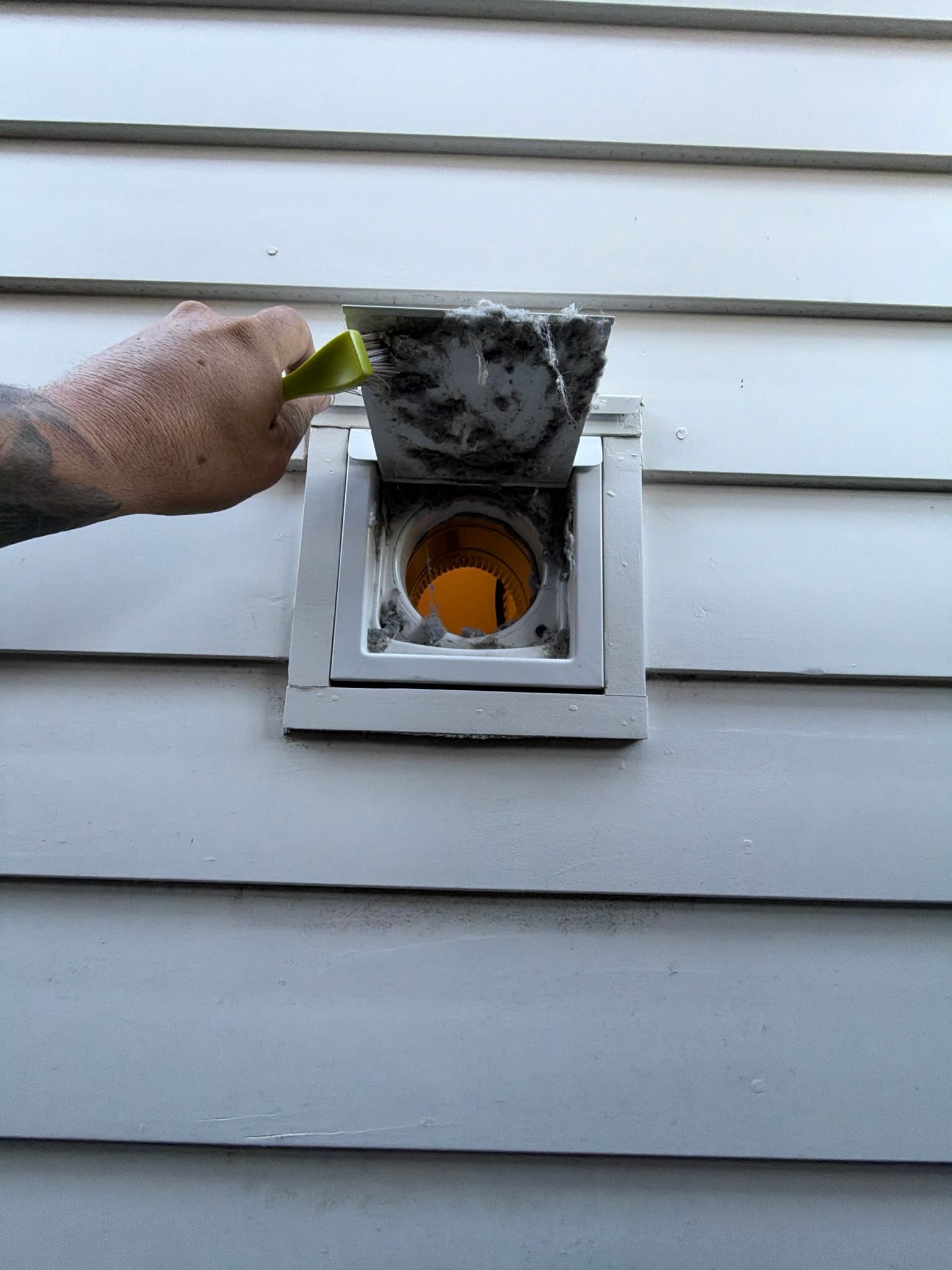 Technician using a hand brush to clear heavy caked lint from the hinged flap and opening of an exterior dryer vent